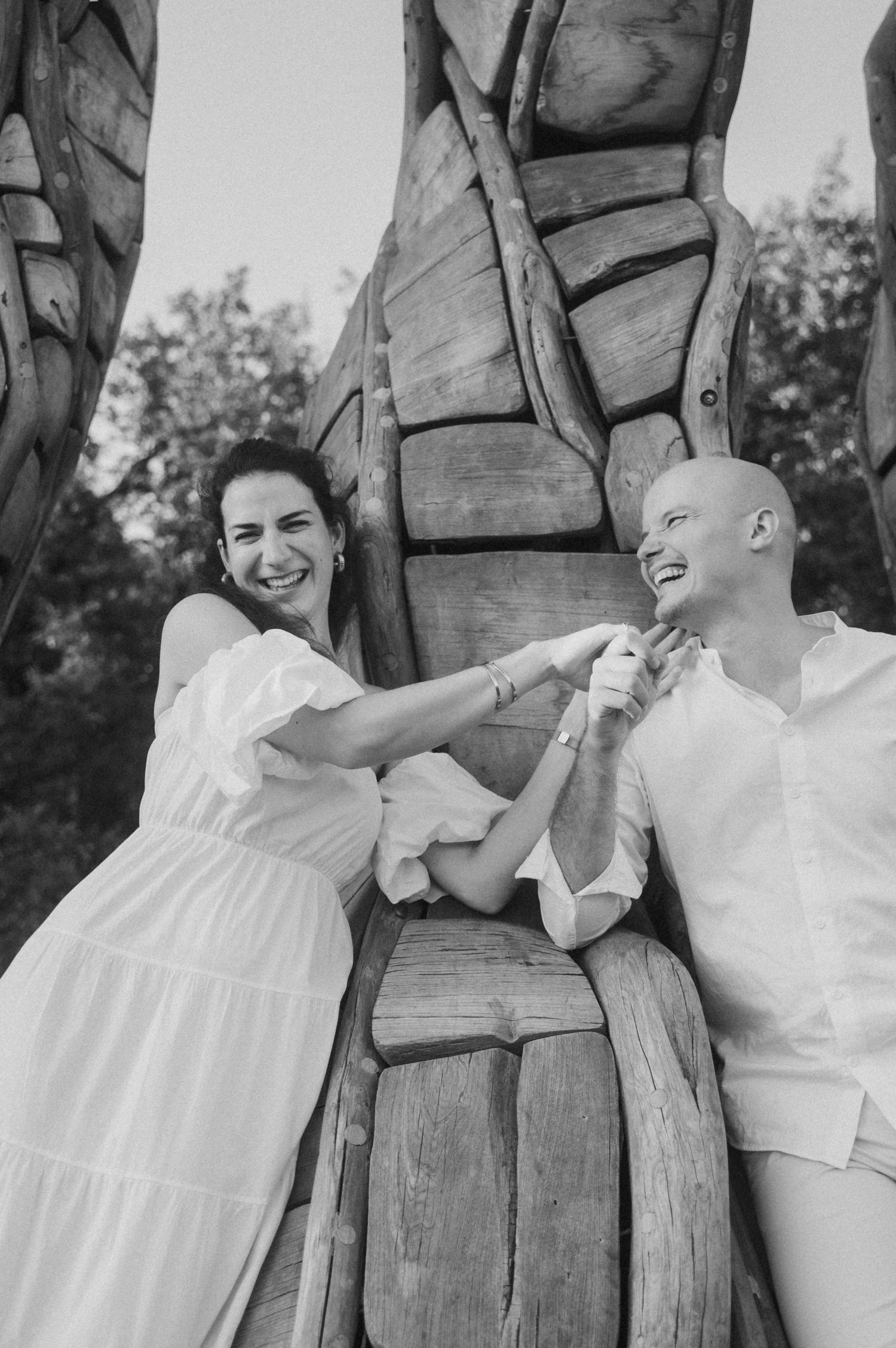 A joyful black and white photo of a couple in white outfits laughing together against a wooden sculpture backdrop.