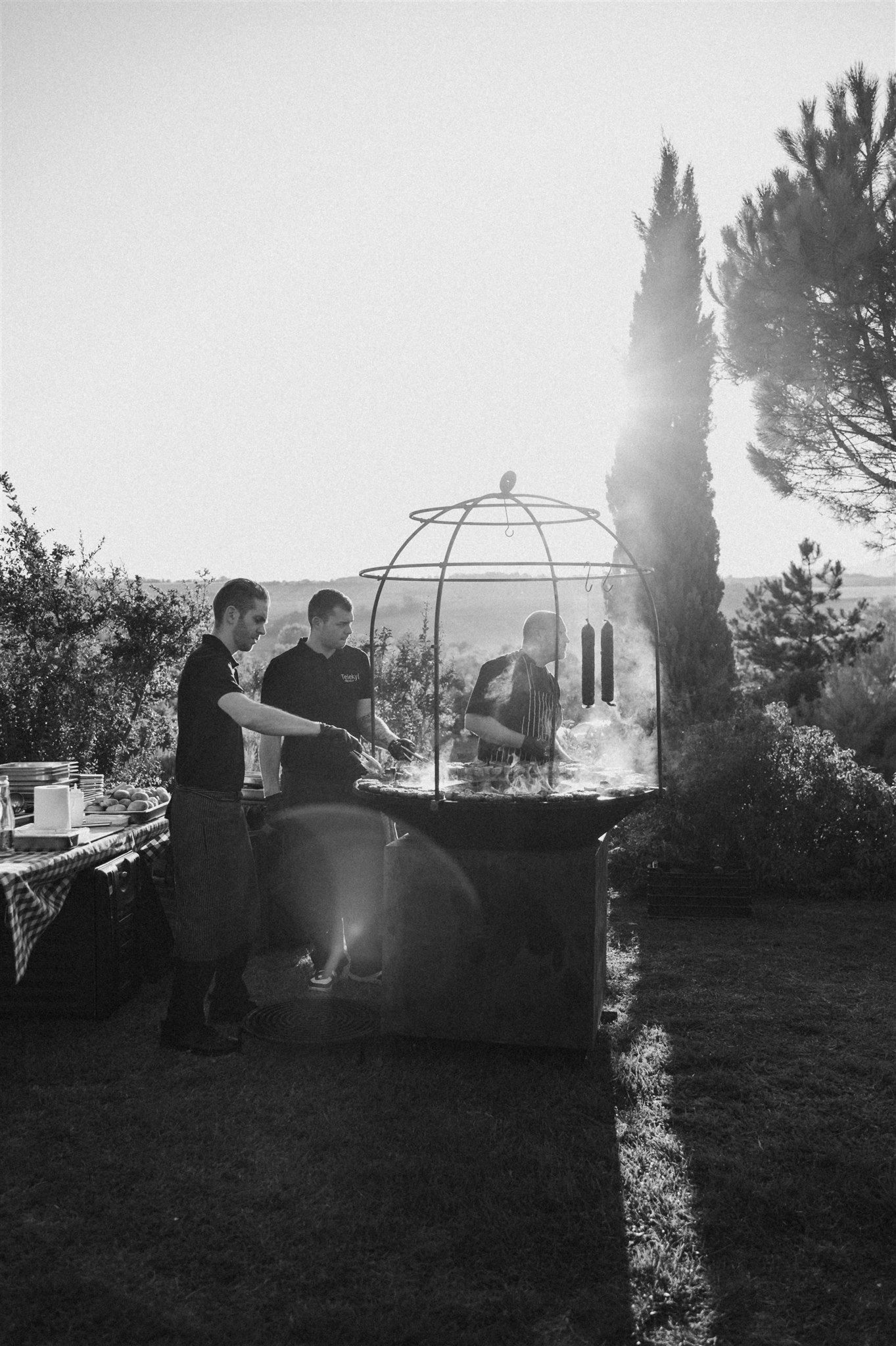 Silhouettes of catering staff working at outdoor cooking stations against a sunset sky.