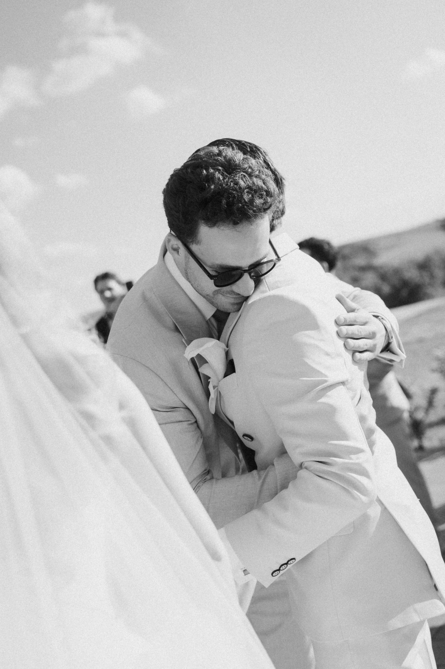 Groom in tan suit and sunglasses sharing an emotional embrace with a guest outdoors on his wedding day.