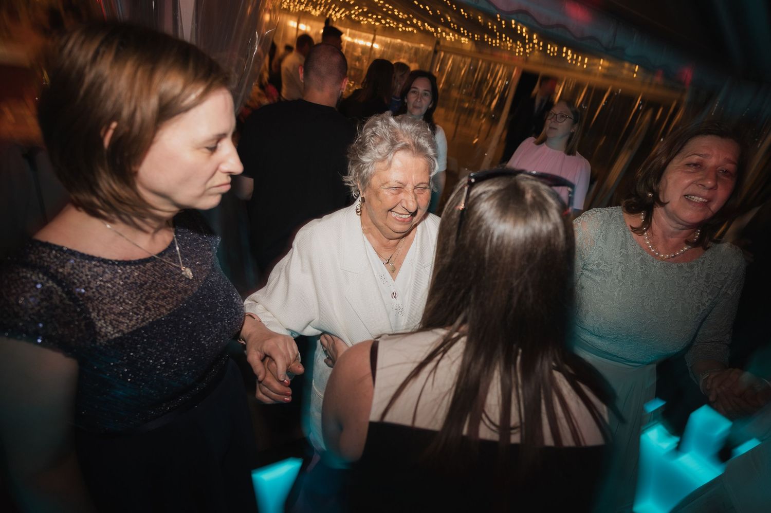 Elderly woman joyfully dancing and holding hands with guests on a glowing wedding reception dance floor.