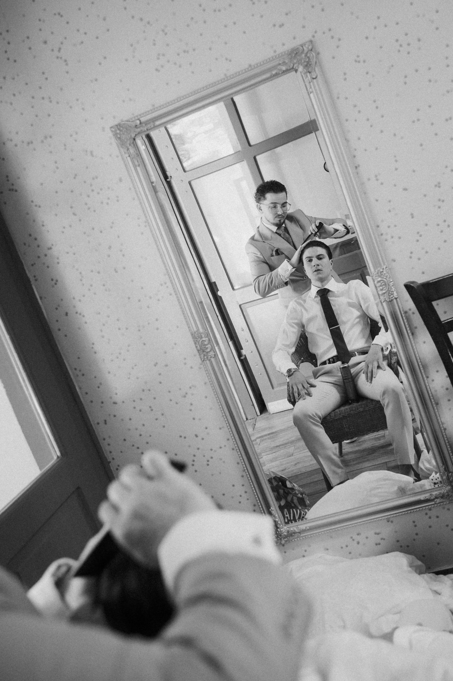 Groom getting ready with groomsman adjusting his tie, reflected in an ornate full-length mirror in black and white.