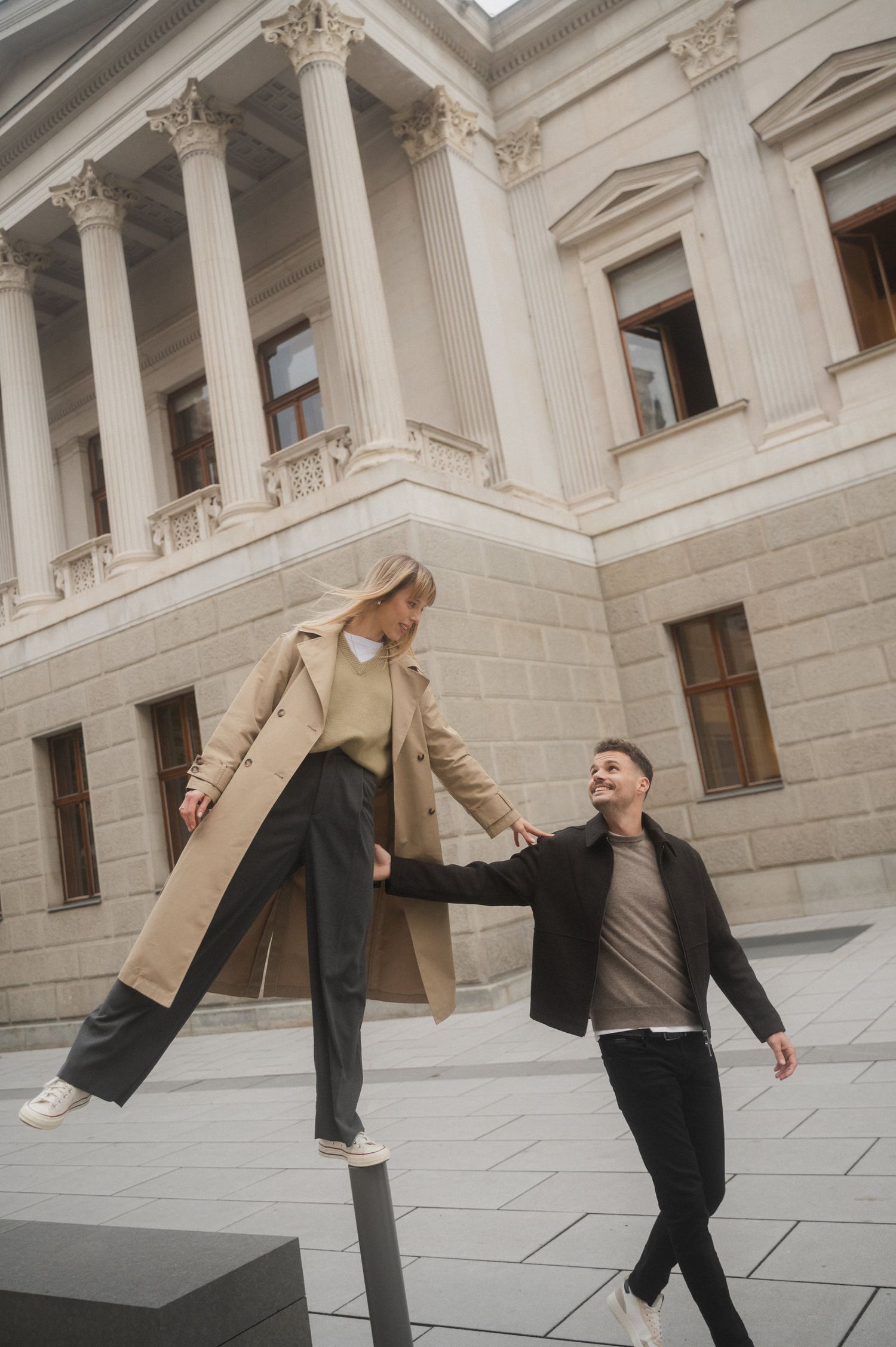 A person in a tan jacket and dark pants performs a sequence of graceful dance movements against a classical stone building.