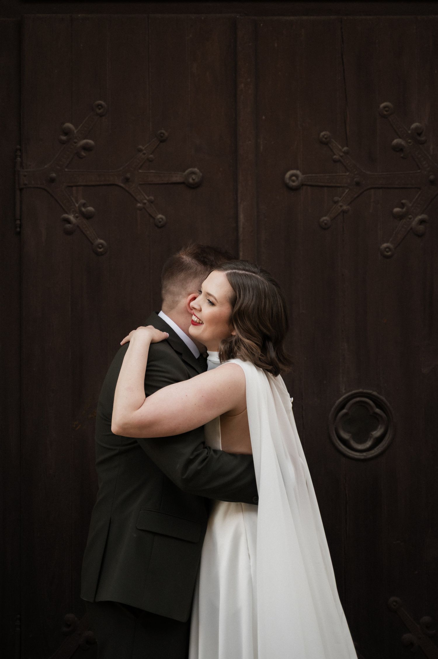 A romantic wedding couple shares an intimate kiss against a dark wooden door with decorative metal details.