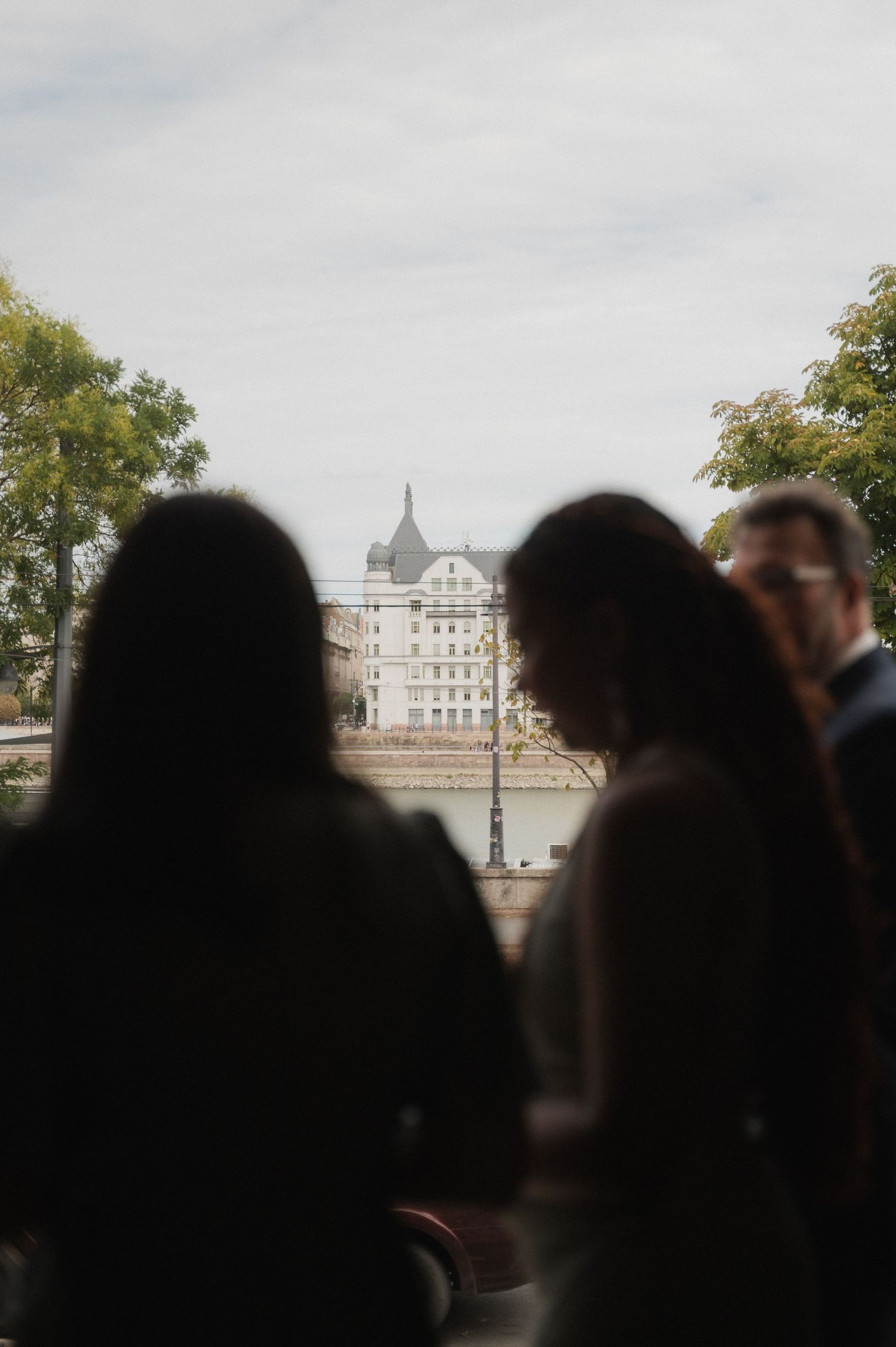 Silhouettes of wedding guests standing before a scenic riverside European cityscape with historic buildings.