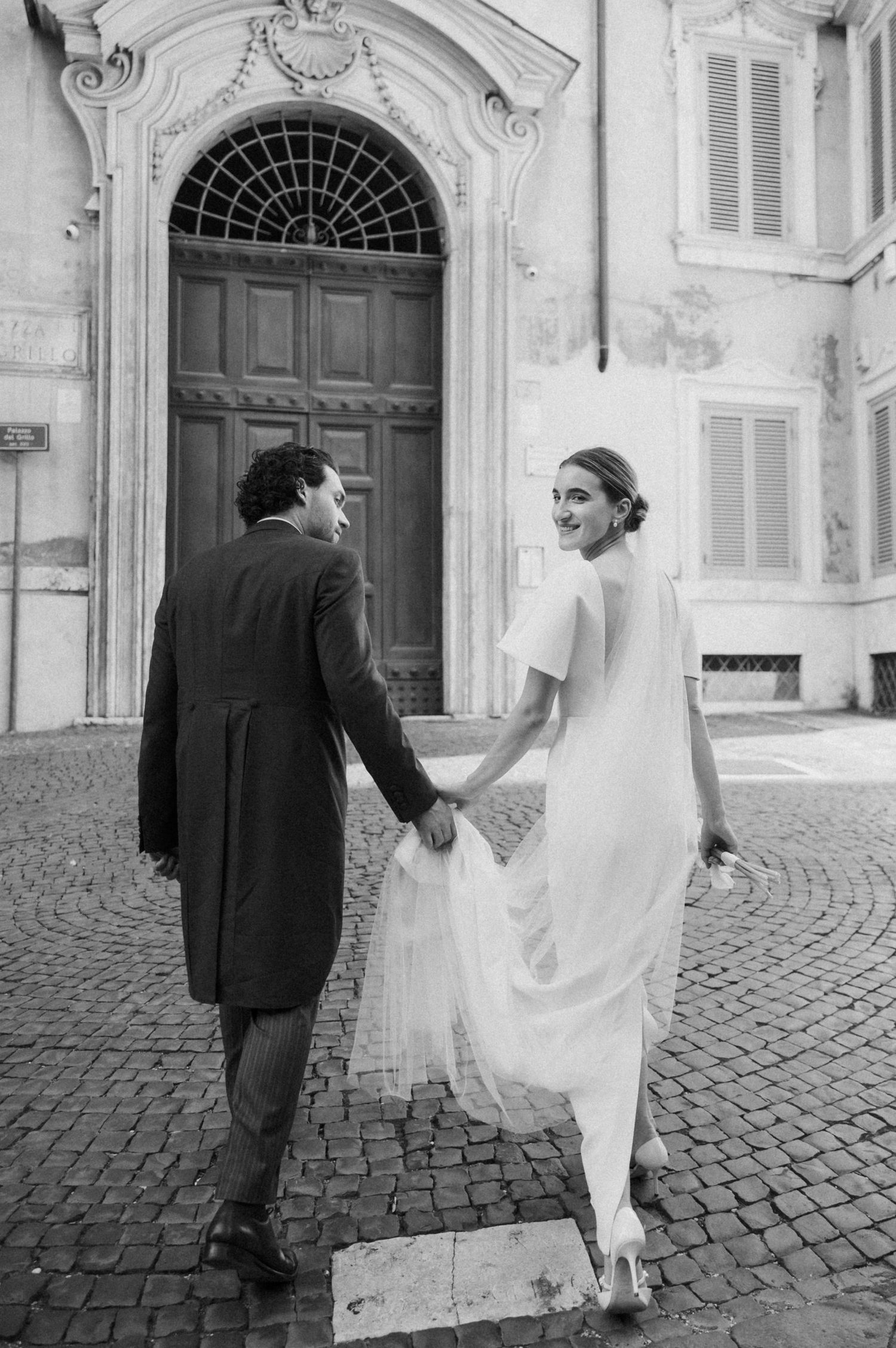 Black and white photo of a bride and groom walking along a European cobblestone street.