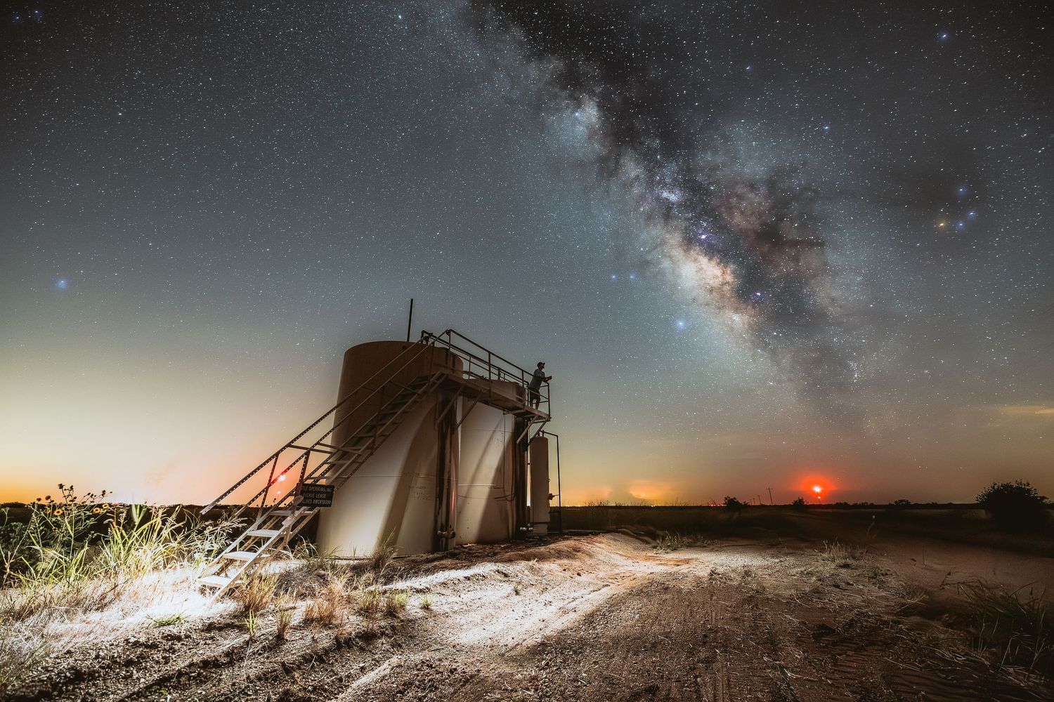 The Milky Way stretches across the night sky above an industrial structure in a desert landscape.