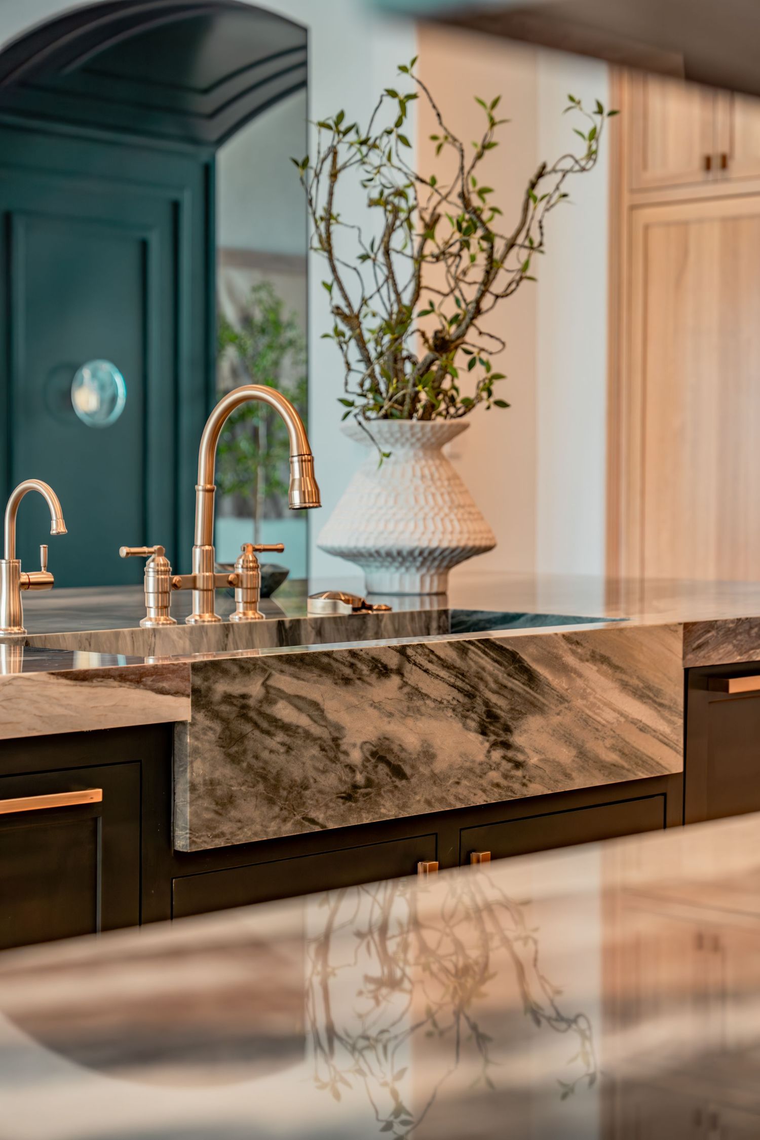 Elegant kitchen detail showing marble farmhouse sink, brass faucets, and decorative white vase with branches.