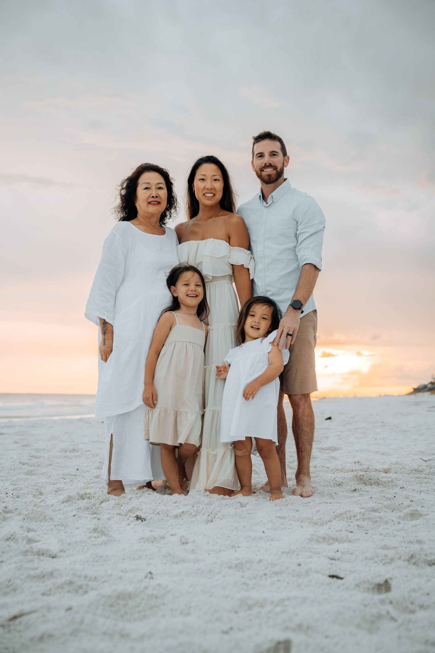 Family wearing white outfits poses together on beach at sunset.