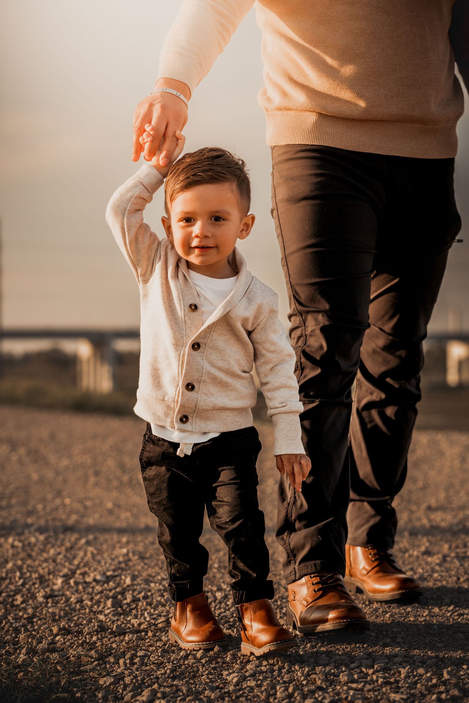 A toddler in white takes first steps with adult guidance on a gravel path.