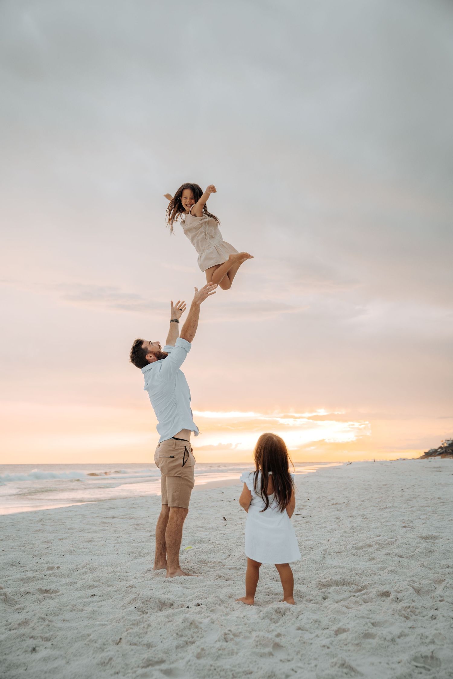 A playful moment captured on a white sandy beach at sunset as someone lifts a child in the air.