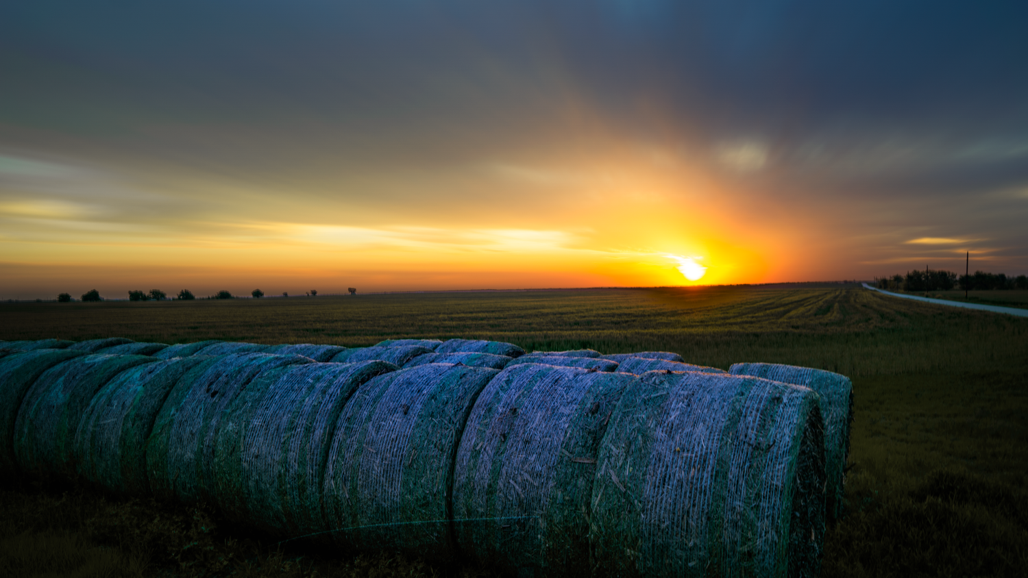 Dramatic sunset over hay bales in a rural field with golden light illuminating the landscape.