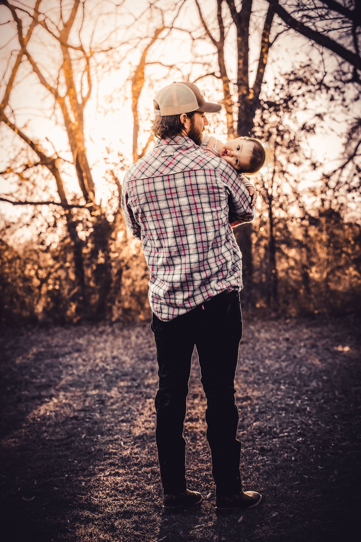 A couple in plaid and denim share an intimate embrace at sunset in a wooded area.