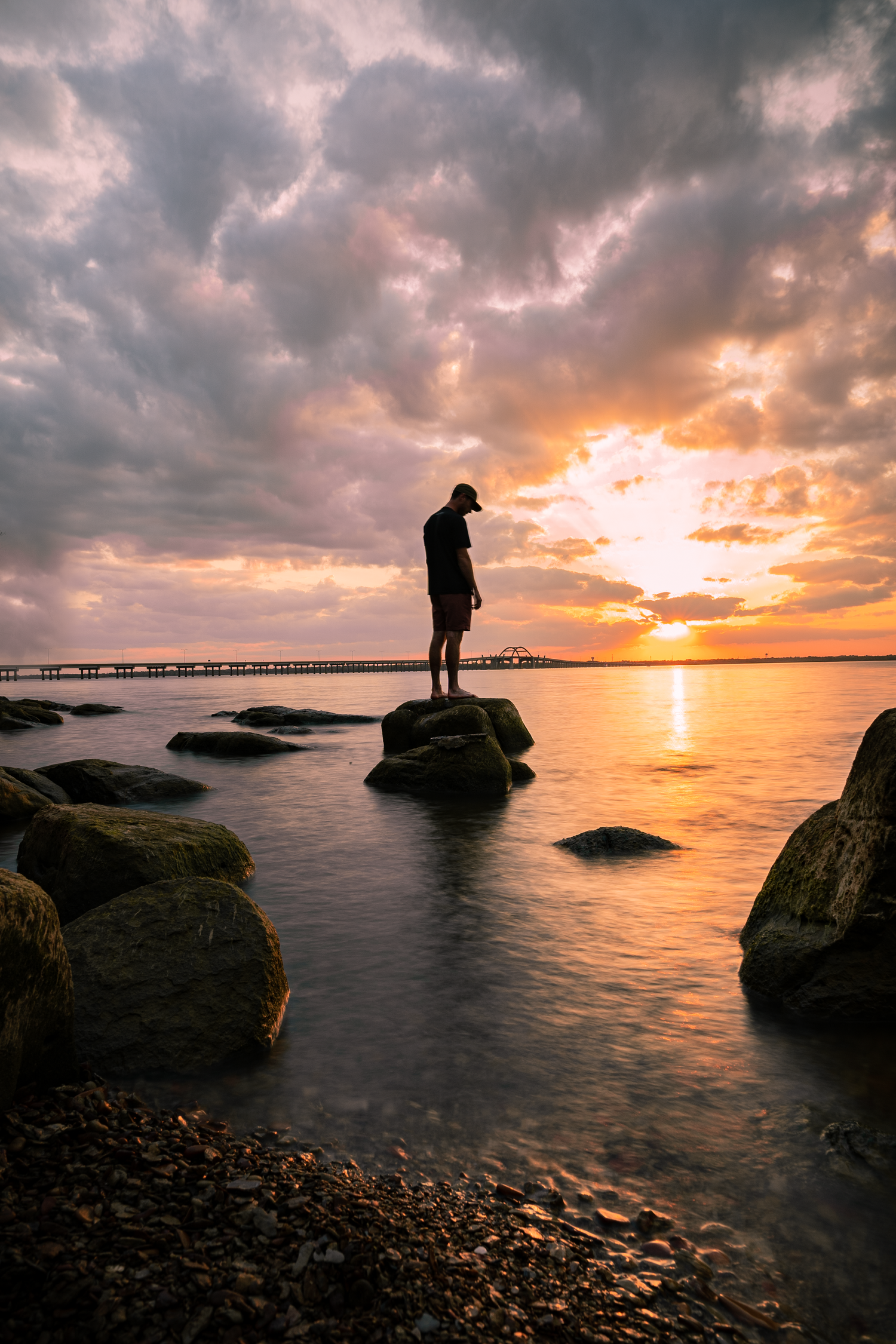Silhouette of person standing on rocks at sunset by ocean pier.