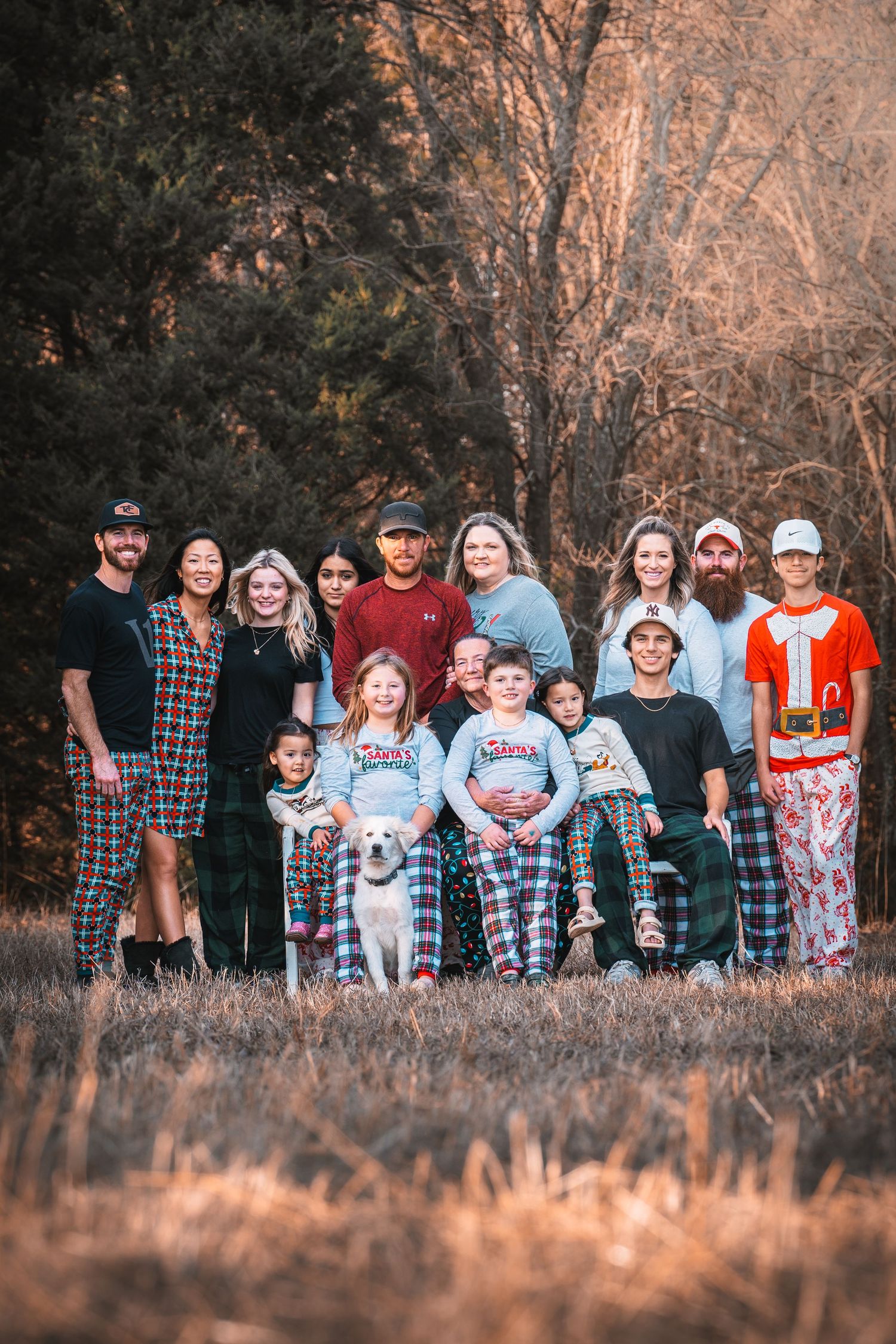 A large family group poses together outdoors at sunset in a field with golden grass and trees in the background. Multigenerational