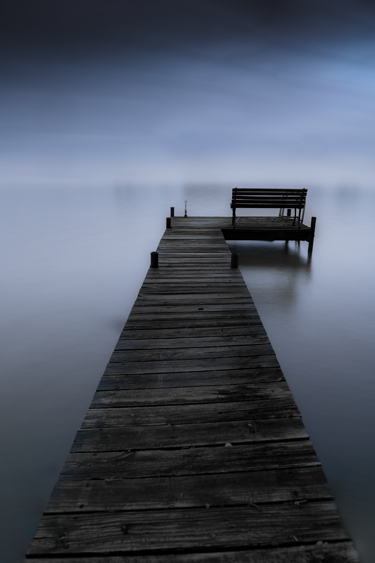 Minimalist wooden pier extending into misty water with lone bench at the end.