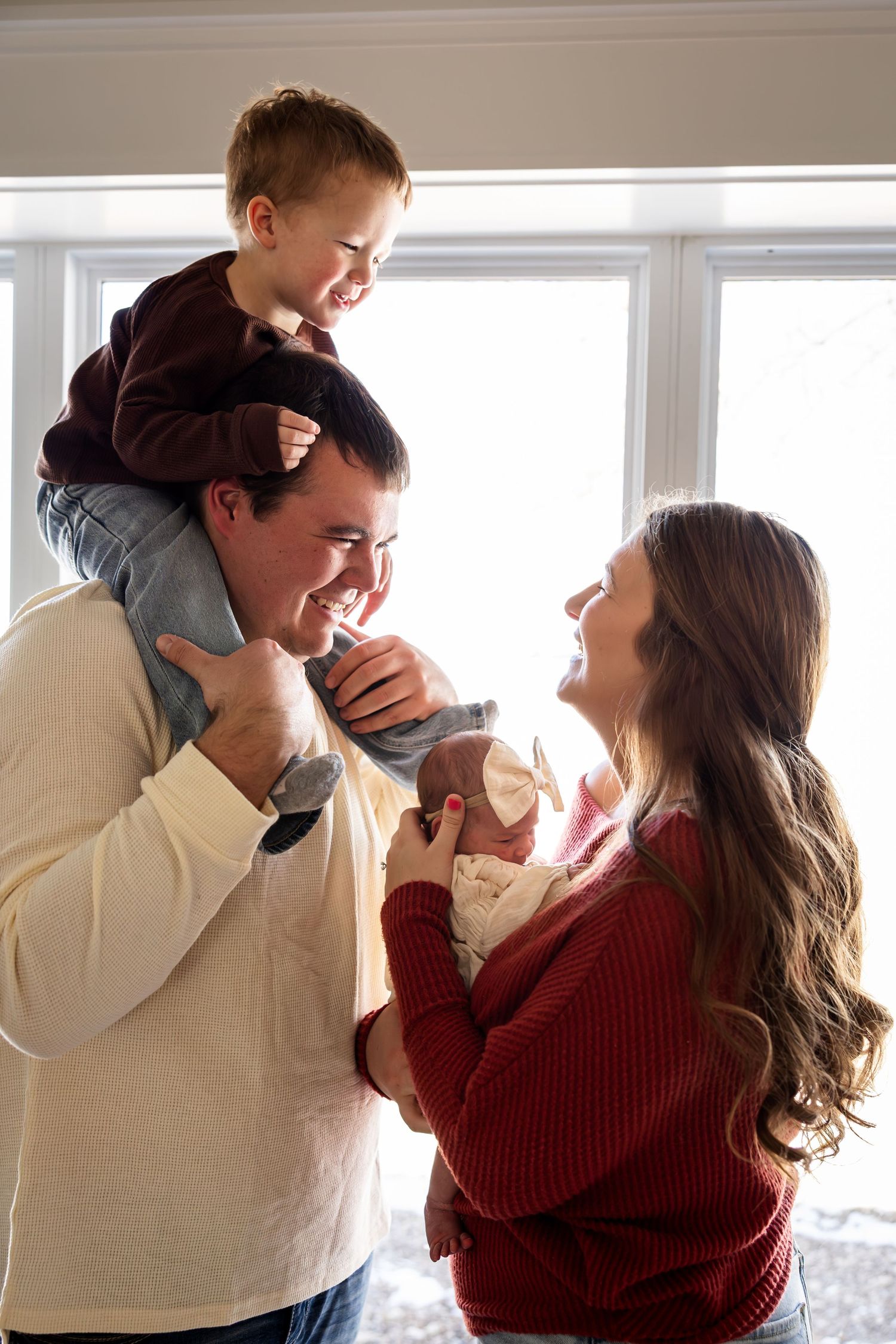 mom holding newborn baby and dad holding toddler on shoulders all smiling and looking at each other during family photos near Macon MO