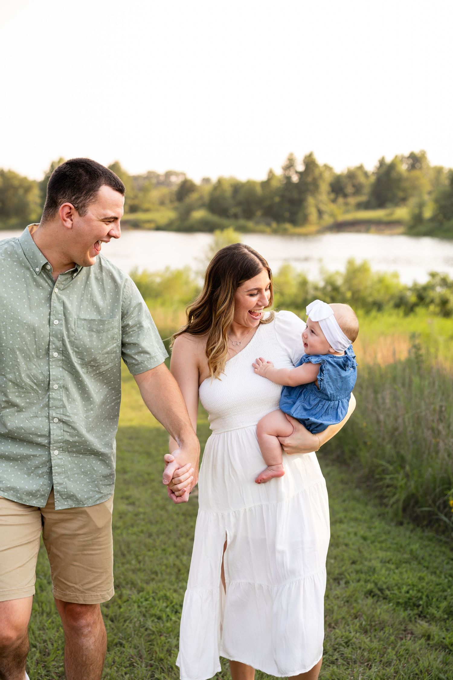 mom and dad holding hands while mom holds infant and smiling at each other while walking with lake in background for family photos in MO