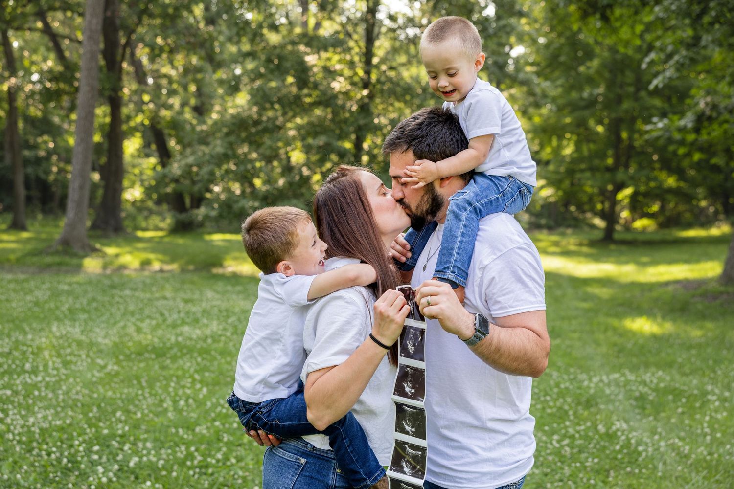 mom and dad kissing while sons are on their back and shoulders holding ultrasound photos for pregnancy announcement family photos at park