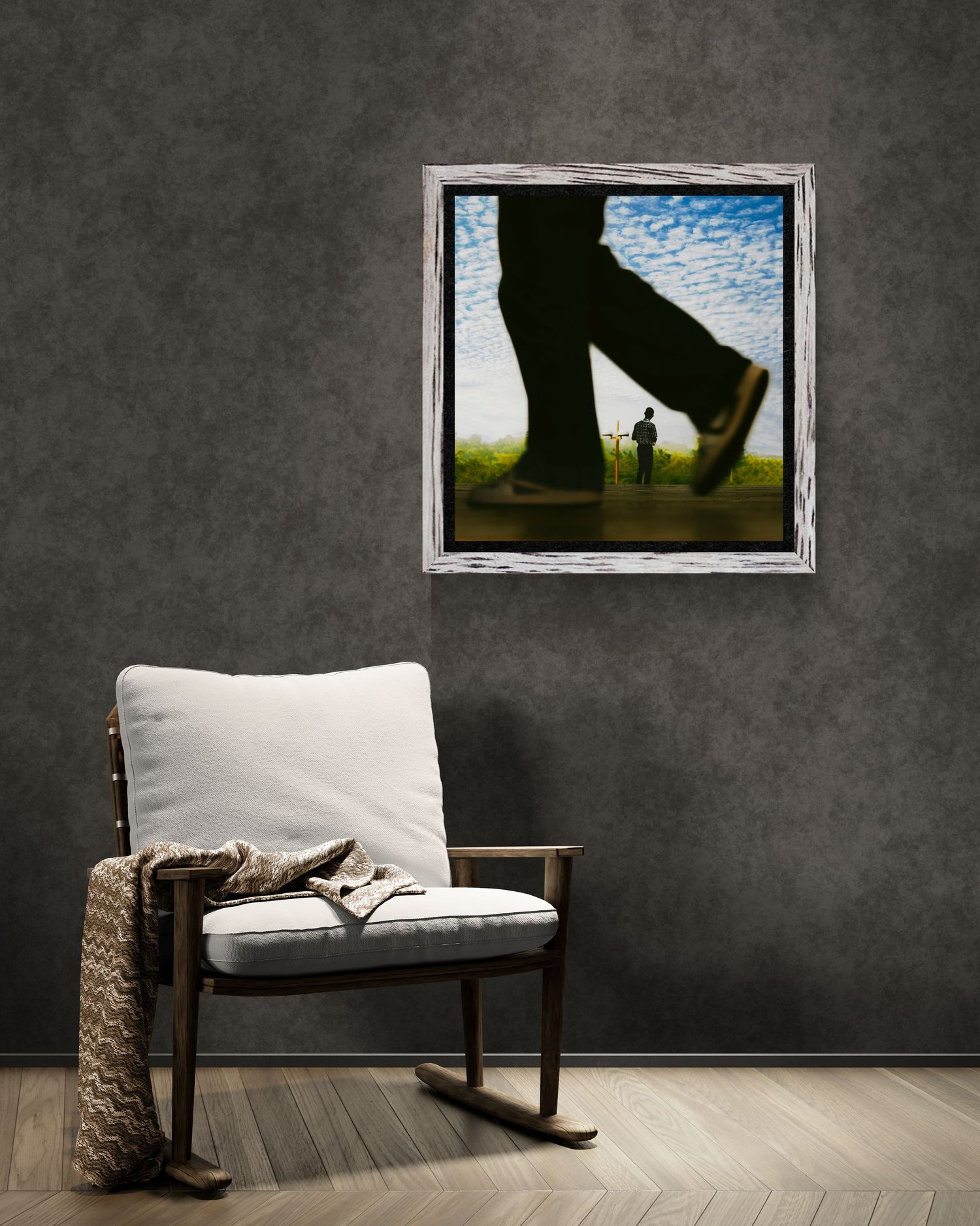 Framed artwork of giant legs walking beside tiny figure on green field, displayed above rocking chair with throw blanket.