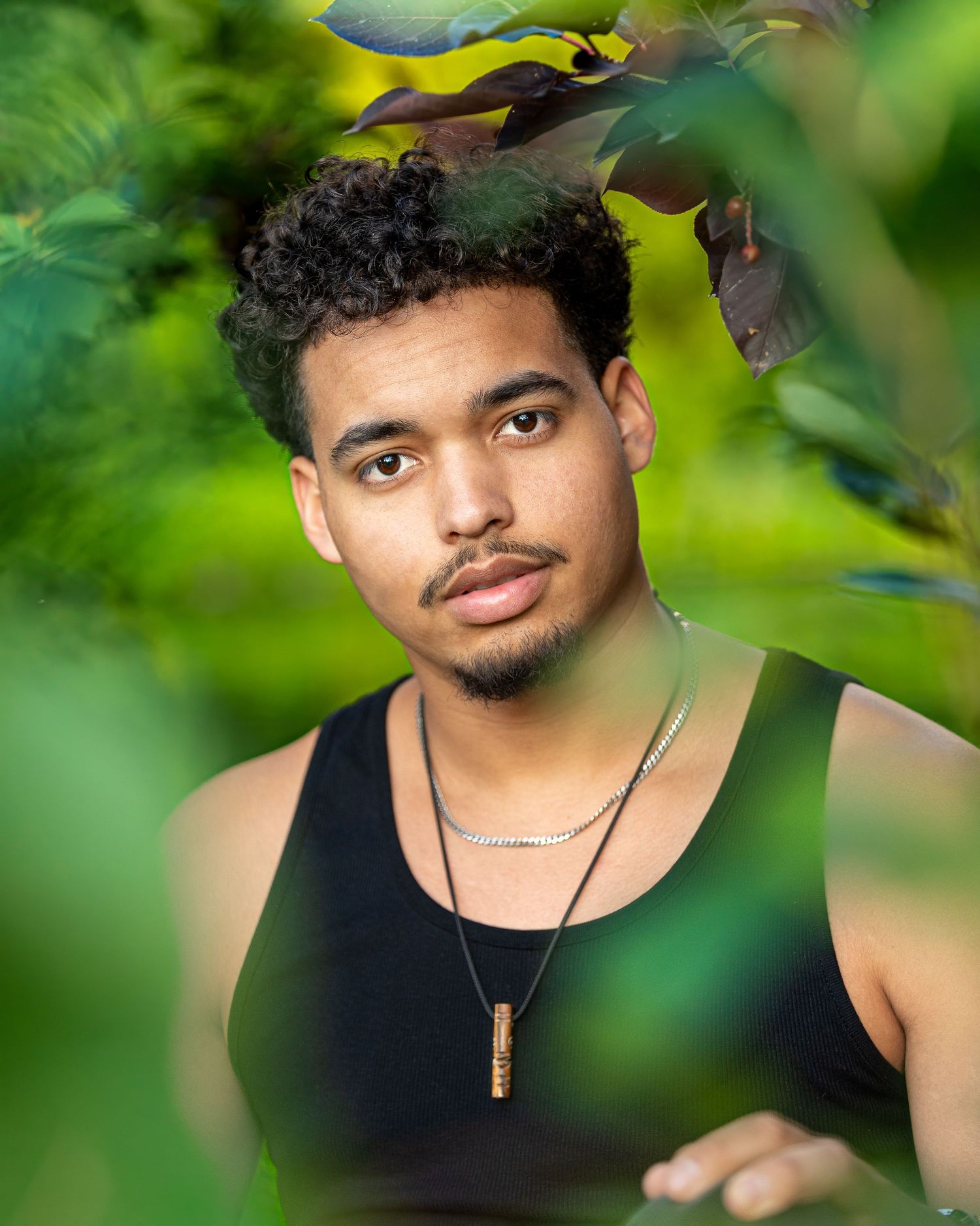Portrait of young man in black tank top with necklace against blurred green foliage background.