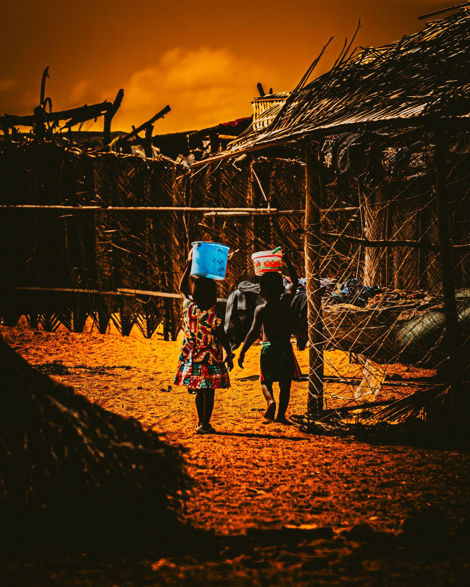 Silhouetted figures walk through a village at sunset casting long shadows on the orange-lit dirt ground.