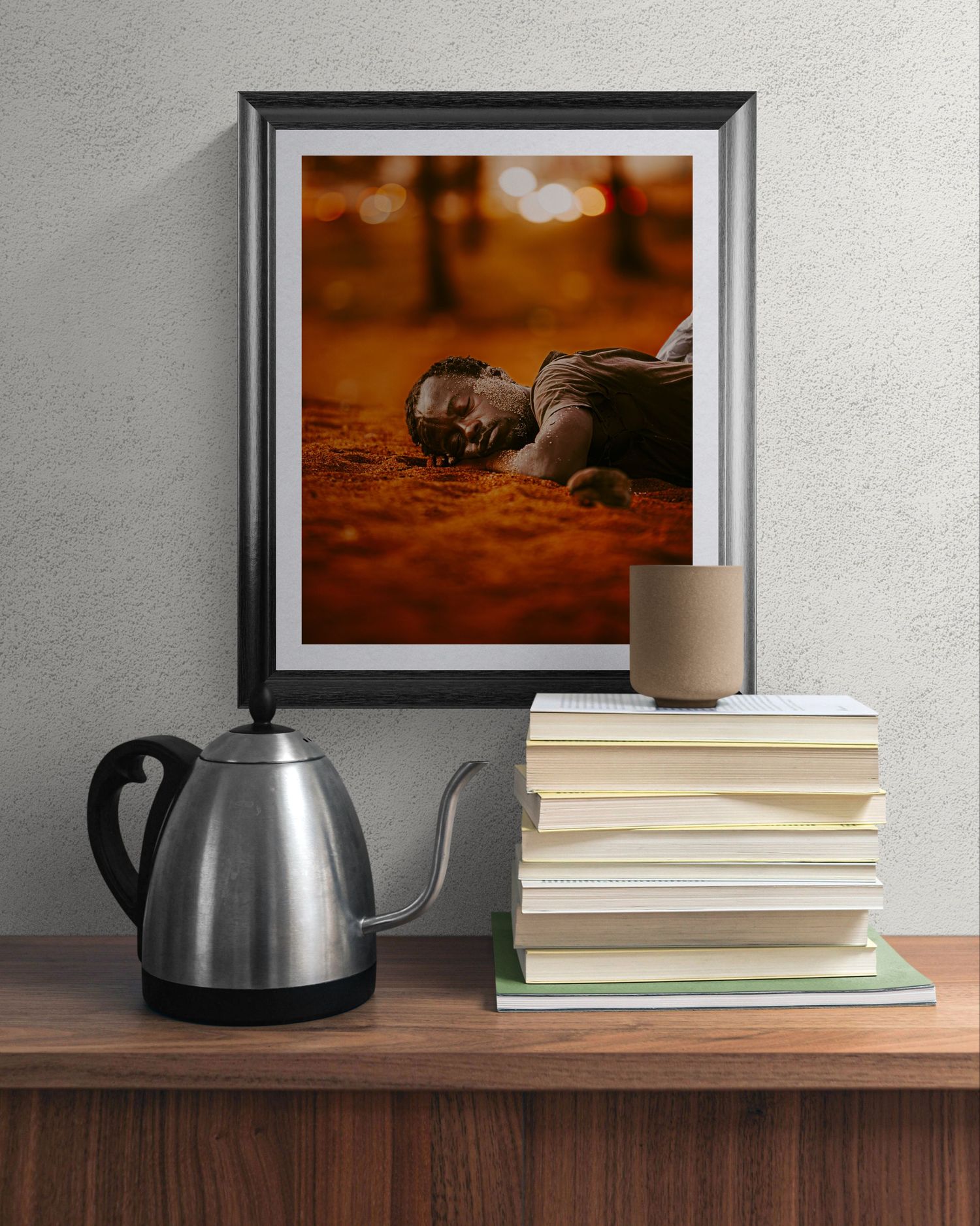 Framed photo of person lying on ground displayed above wooden shelf with steel kettle and stacked books.