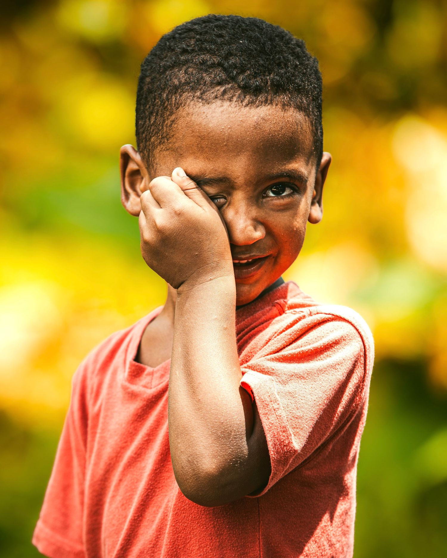 A young boy in a red shirt covers his eye with his hand, smiling shyly outdoors with a blurred green background.