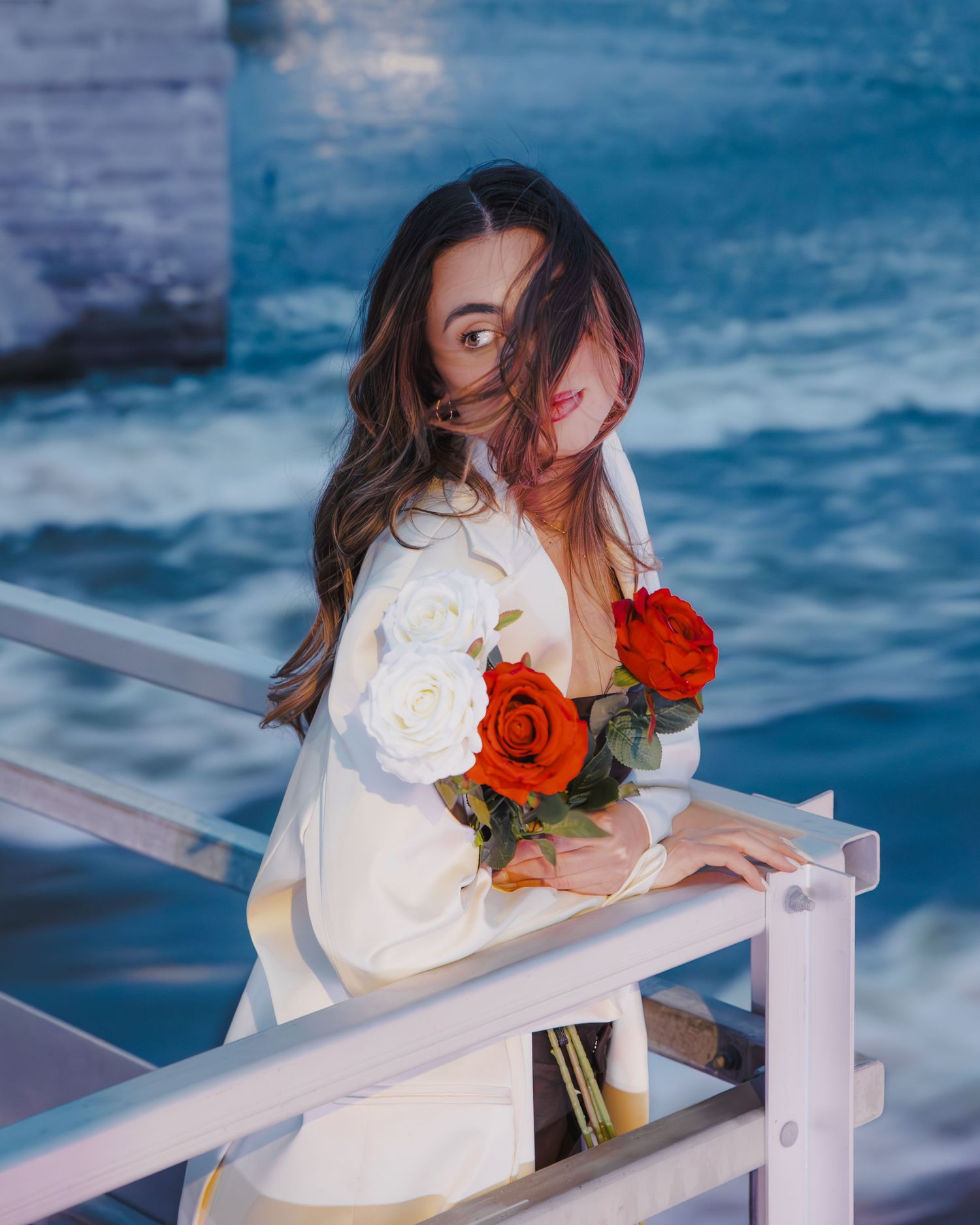 Woman in white outfit holding red and white roses leans on railing with ocean waves in background.