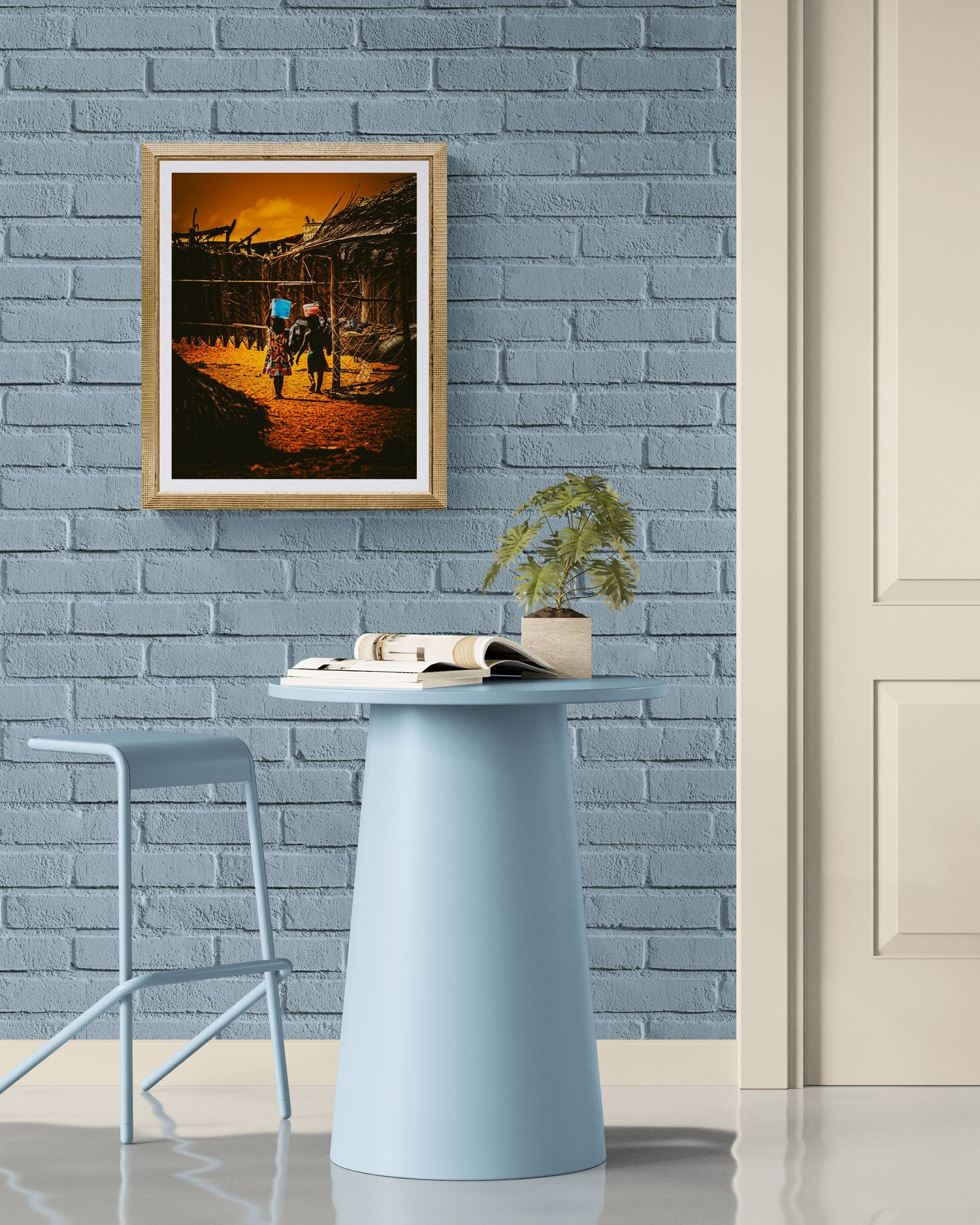 Blue pedestal table and chair against a painted brick wall with framed artwork above, creating a modern minimalist workspace.