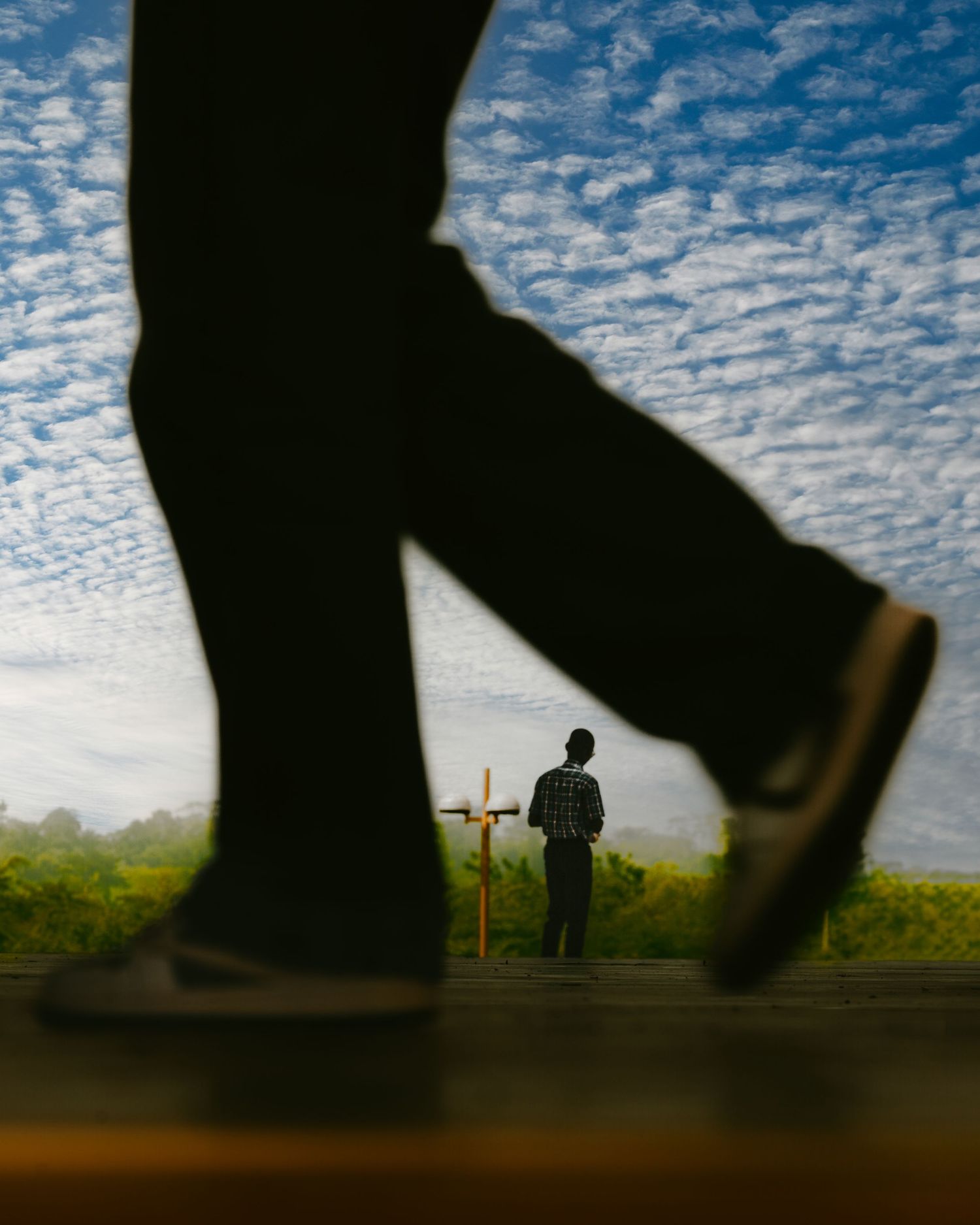 A silhouetted figure stands beneath a dramatic cloudy sky while walking feet create shadows in the foreground.
