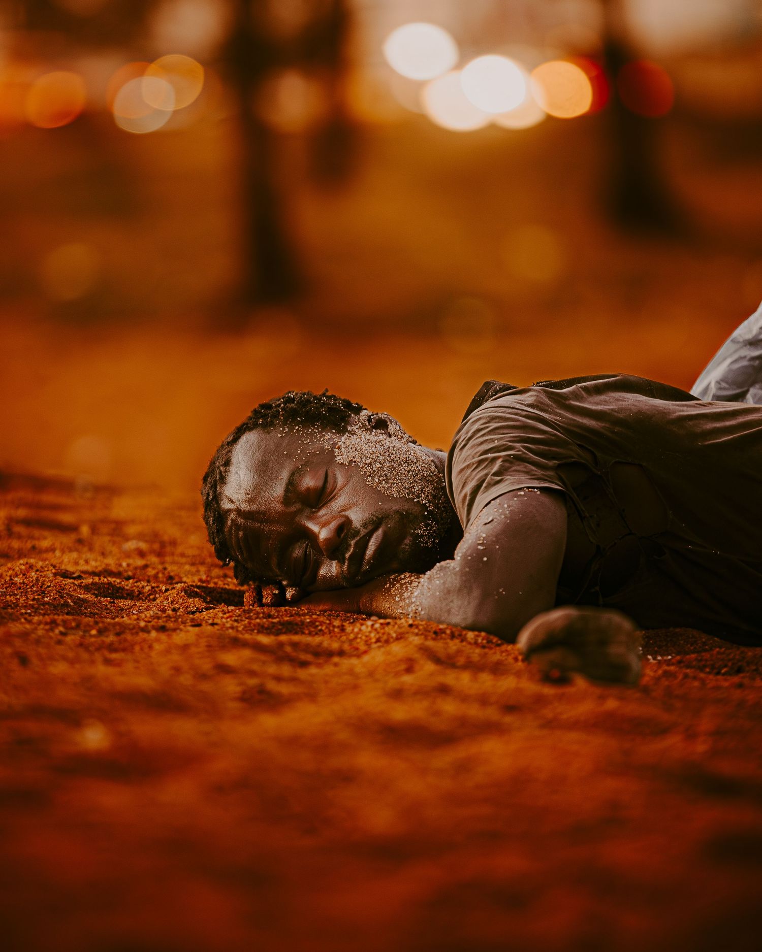 Person lying peacefully on ground covered in warm orange autumn leaves at sunset with bokeh lights in background.