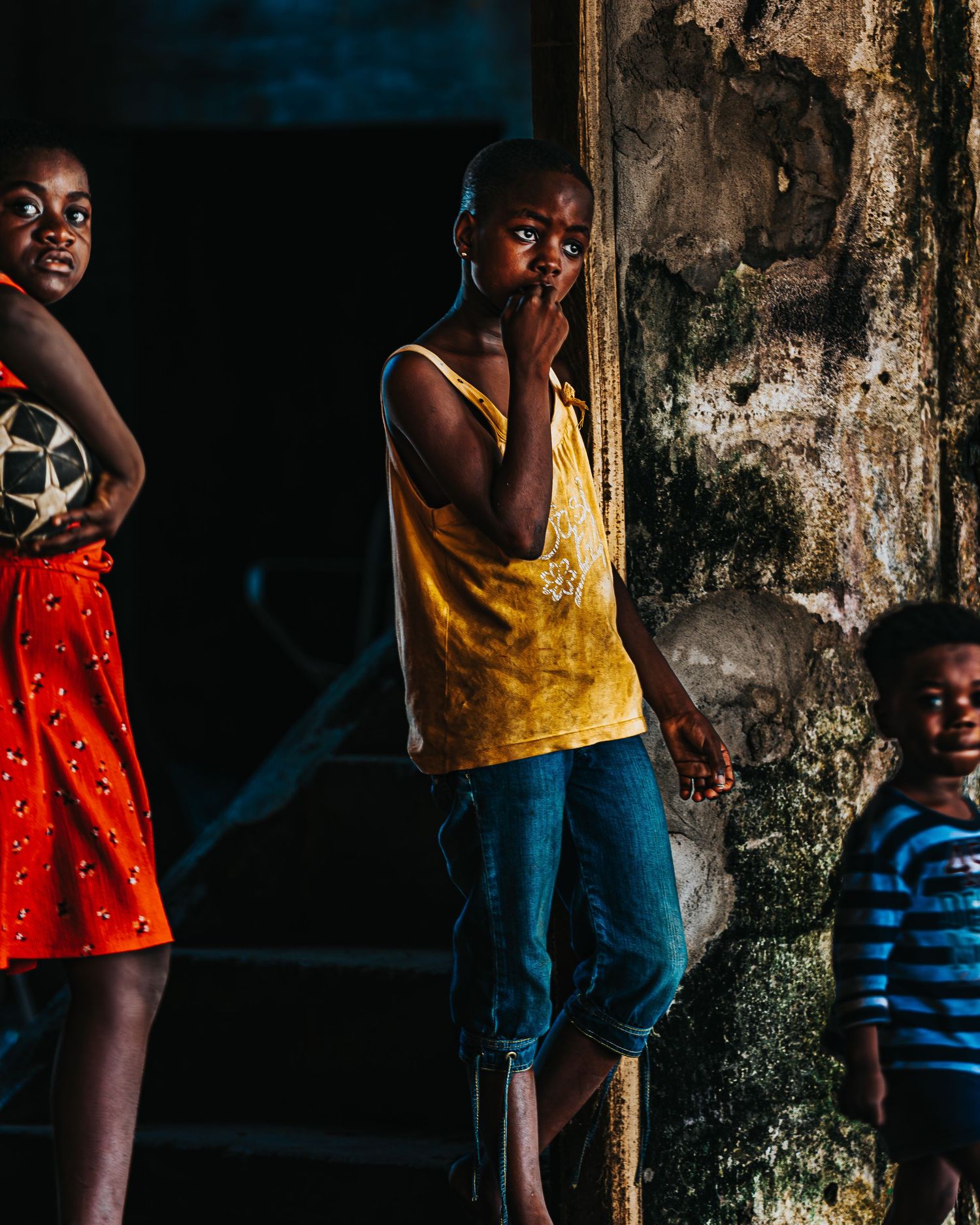 Three African children standing near a weathered wall, one holding a soccer ball, another in a yellow tank top.