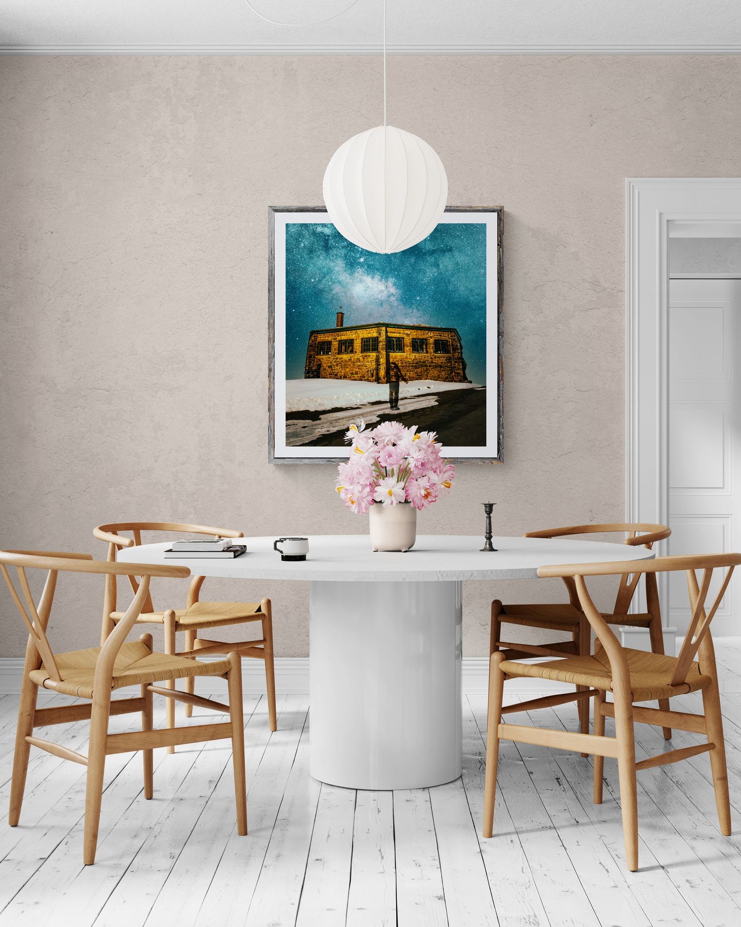 Minimalist dining room with white oval table, wooden Wishbone chairs, floral centerpiece, and framed starry night artwork on wall.