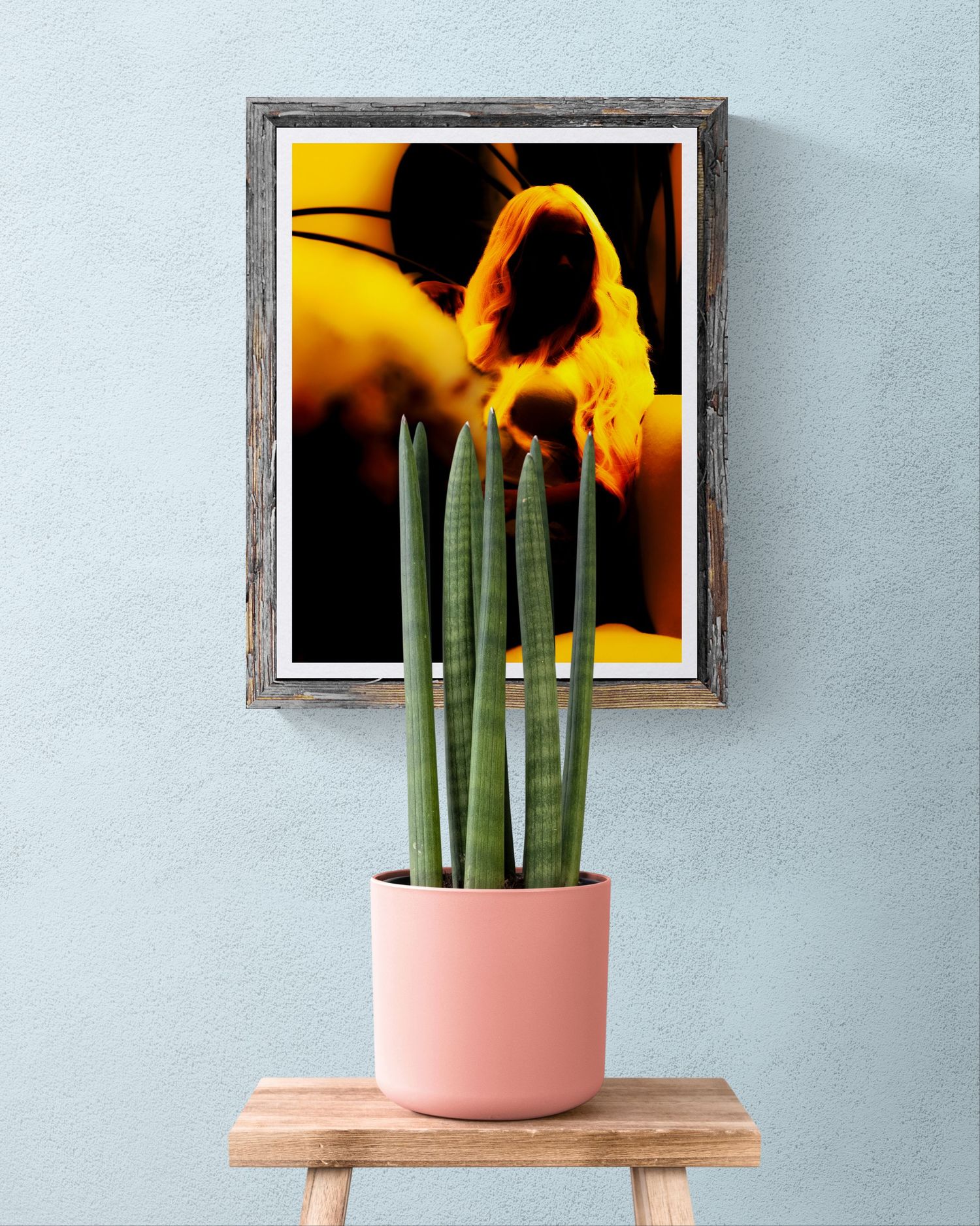 A framed yellow floral abstract art print hangs on a light blue wall above a cactus in a pink pot on a wooden stool.