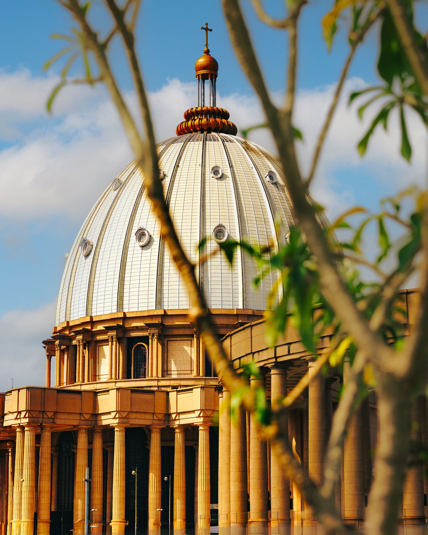 Ornate church dome with golden cross viewed through tree branches against blue sky with white clouds.