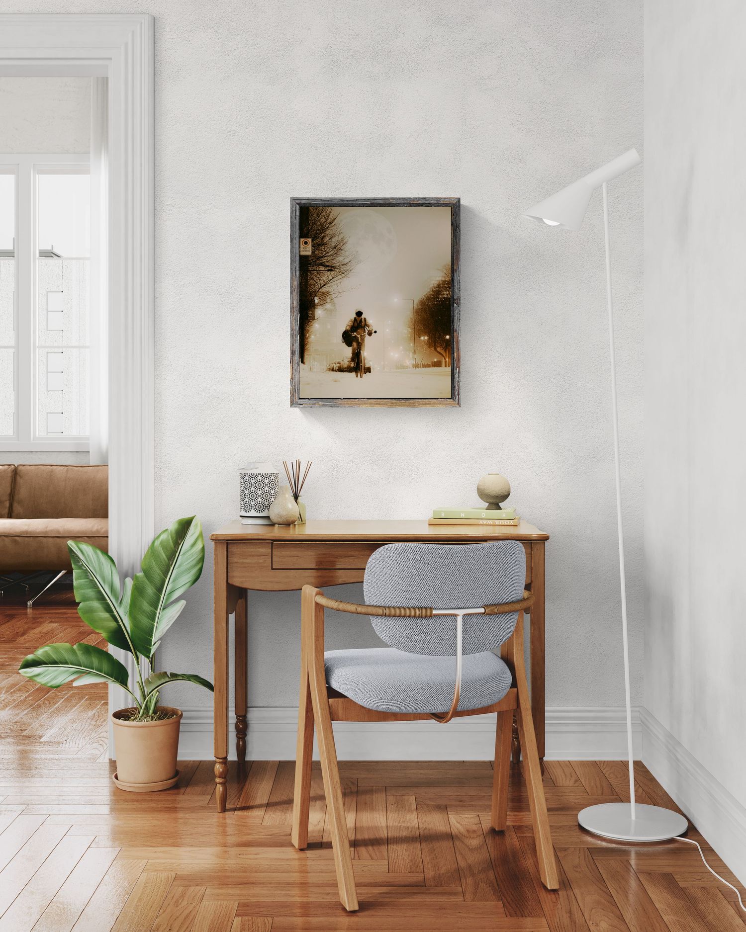 Minimalist home office with wooden desk, gray chair, floor lamp, potted plant, and framed winter artwork on white wall.