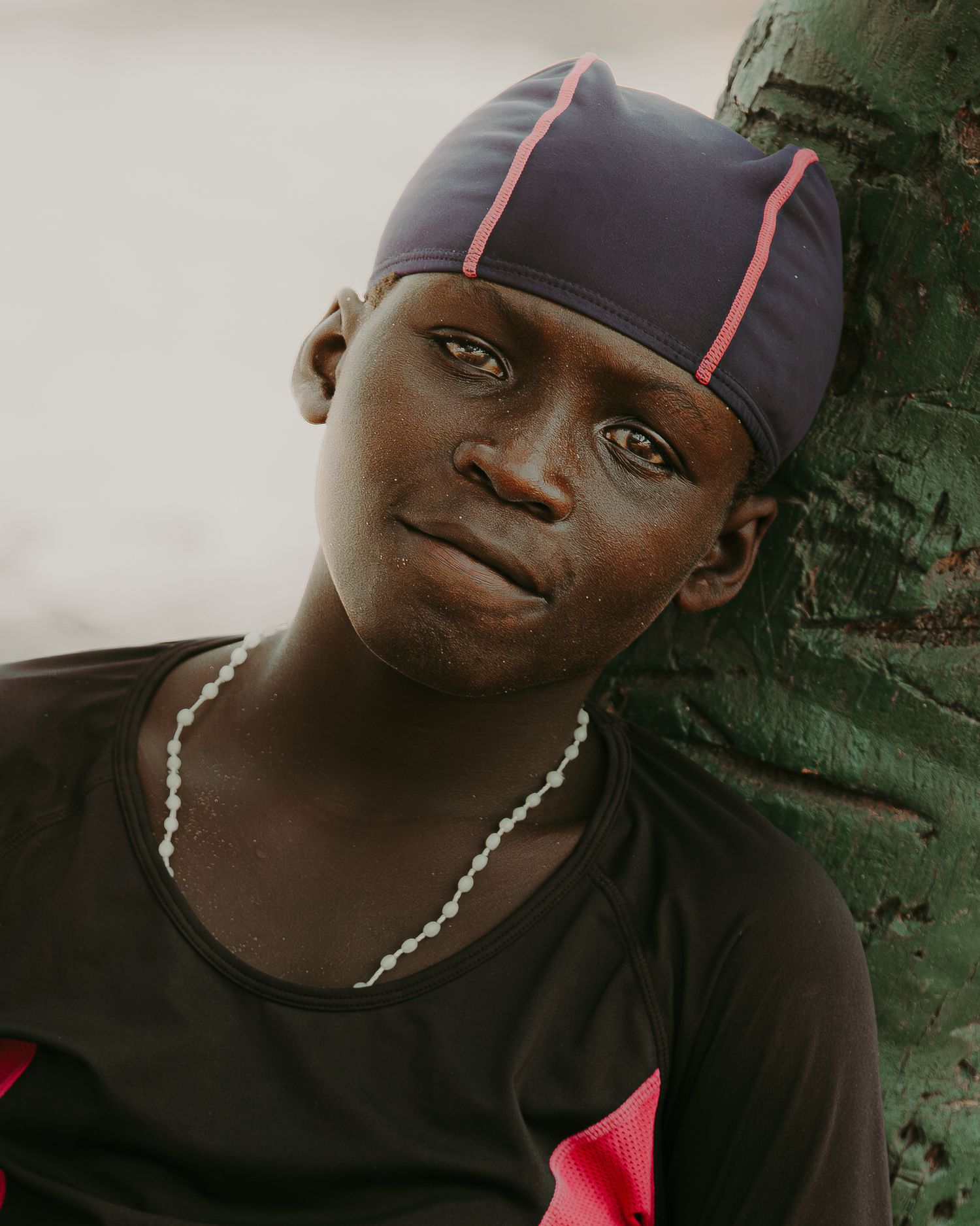 Young boy wearing a dark blue du-rag and black athletic shirt, leaning against a tree trunk.