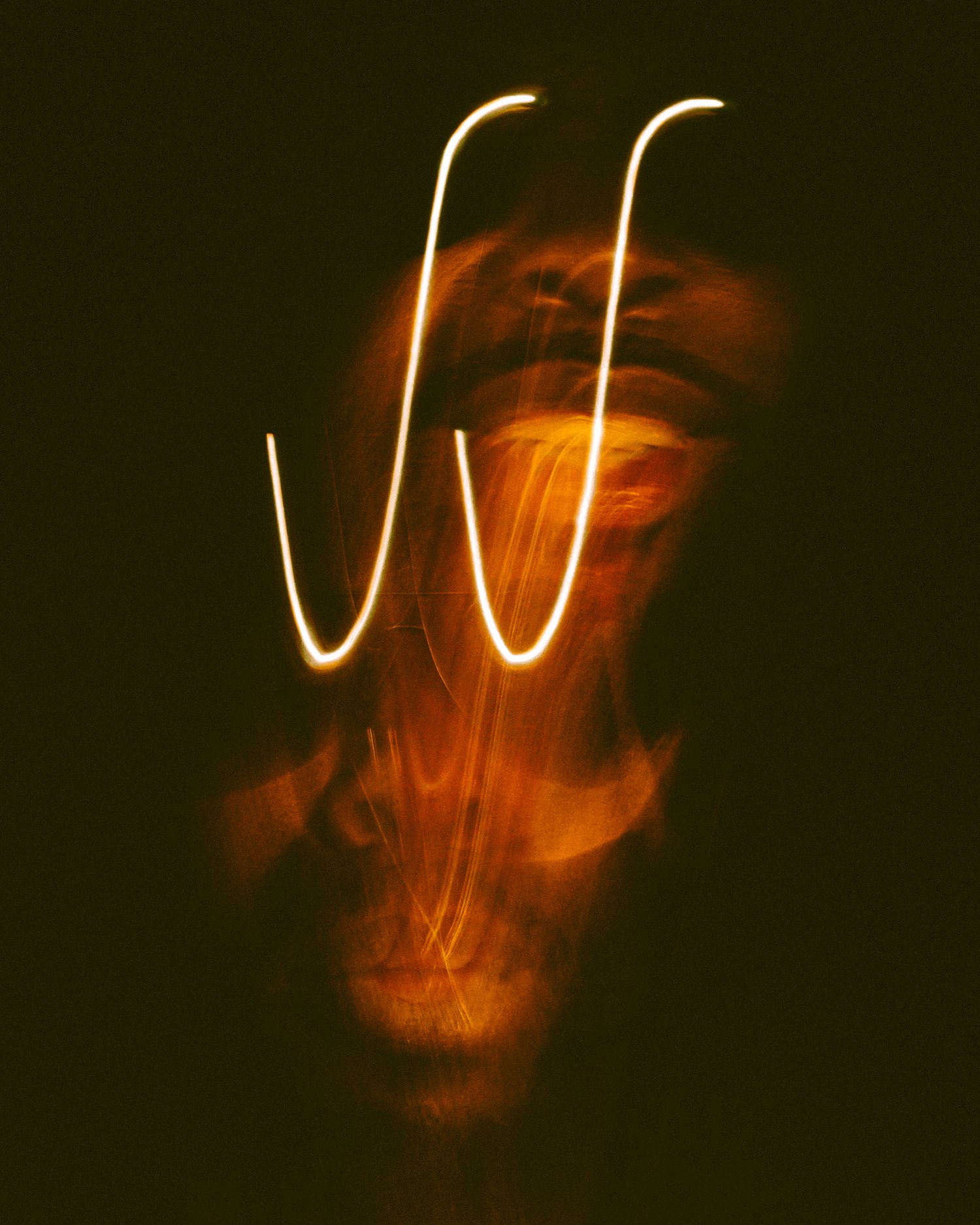 Long exposure light painting creates glowing orange mask-like patterns against a dark background.