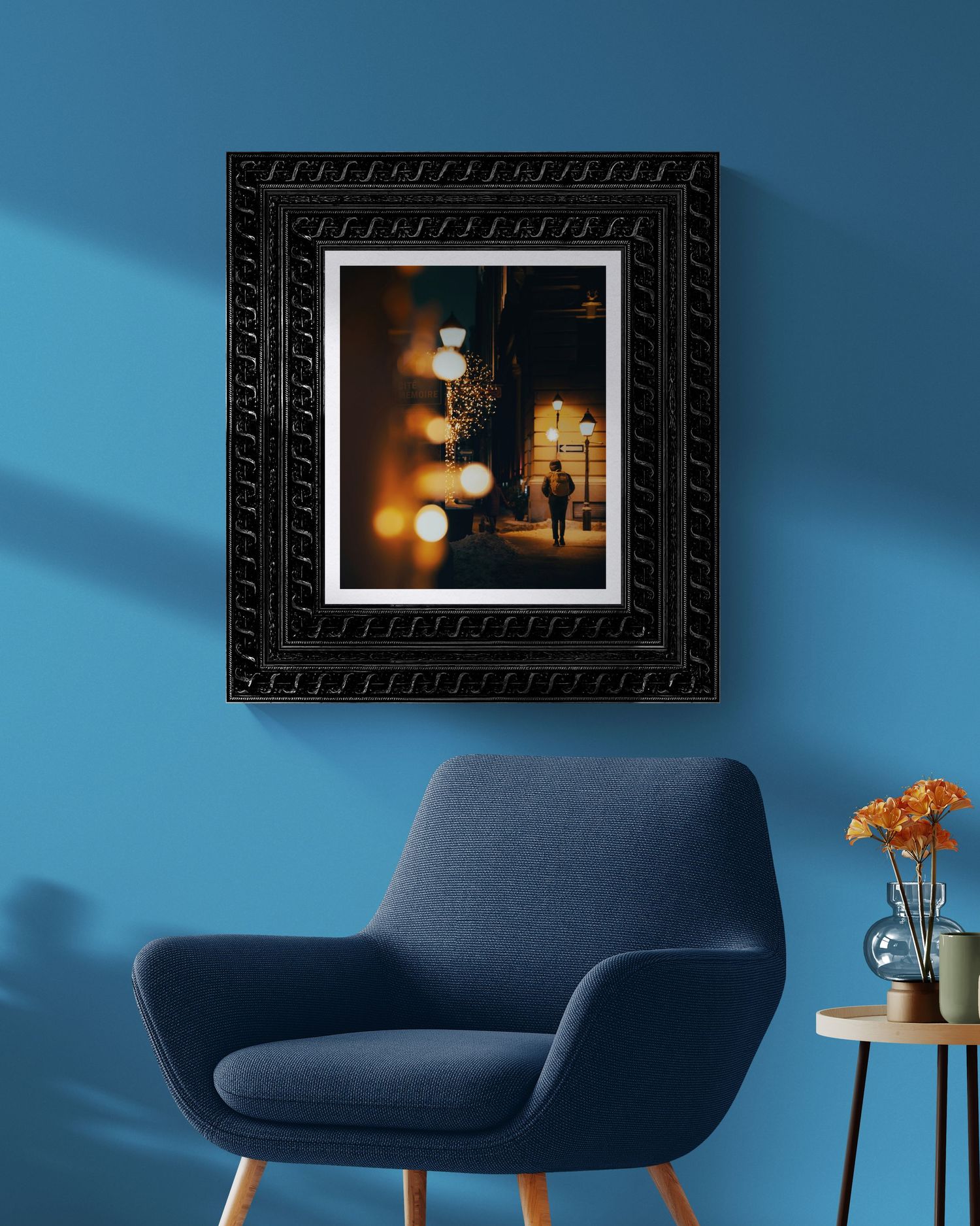 Framed night street photography print in ornate black frame on blue wall above modern navy armchair with side table.