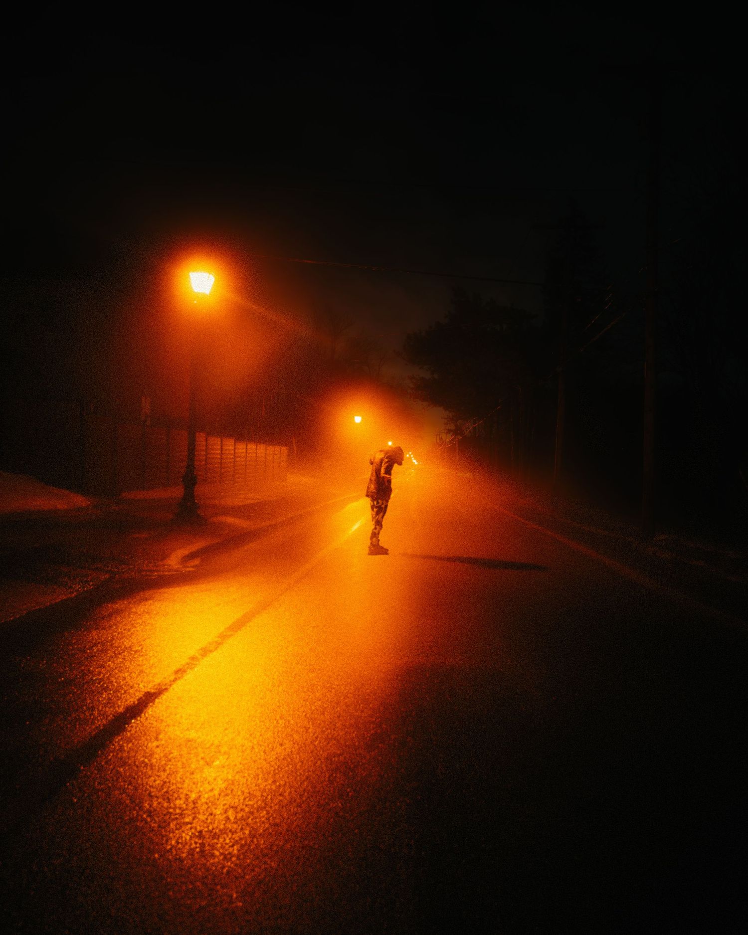 A lone figure walks on a foggy night street illuminated by warm orange street lamps reflecting on wet pavement.