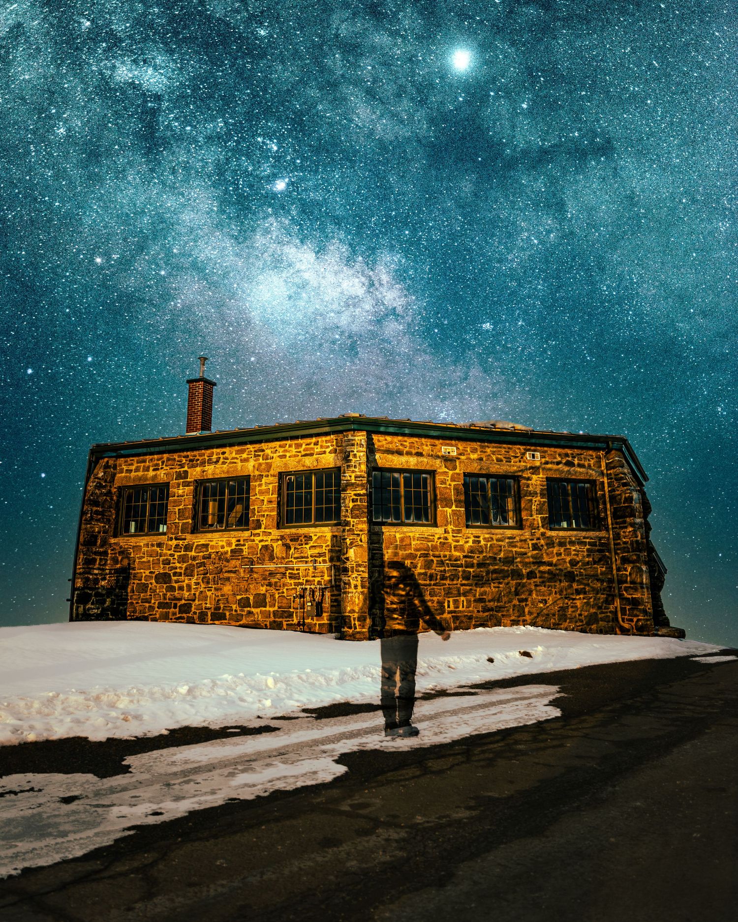 Person standing before a stone building under a stunning Milky Way night sky with snow on the ground.