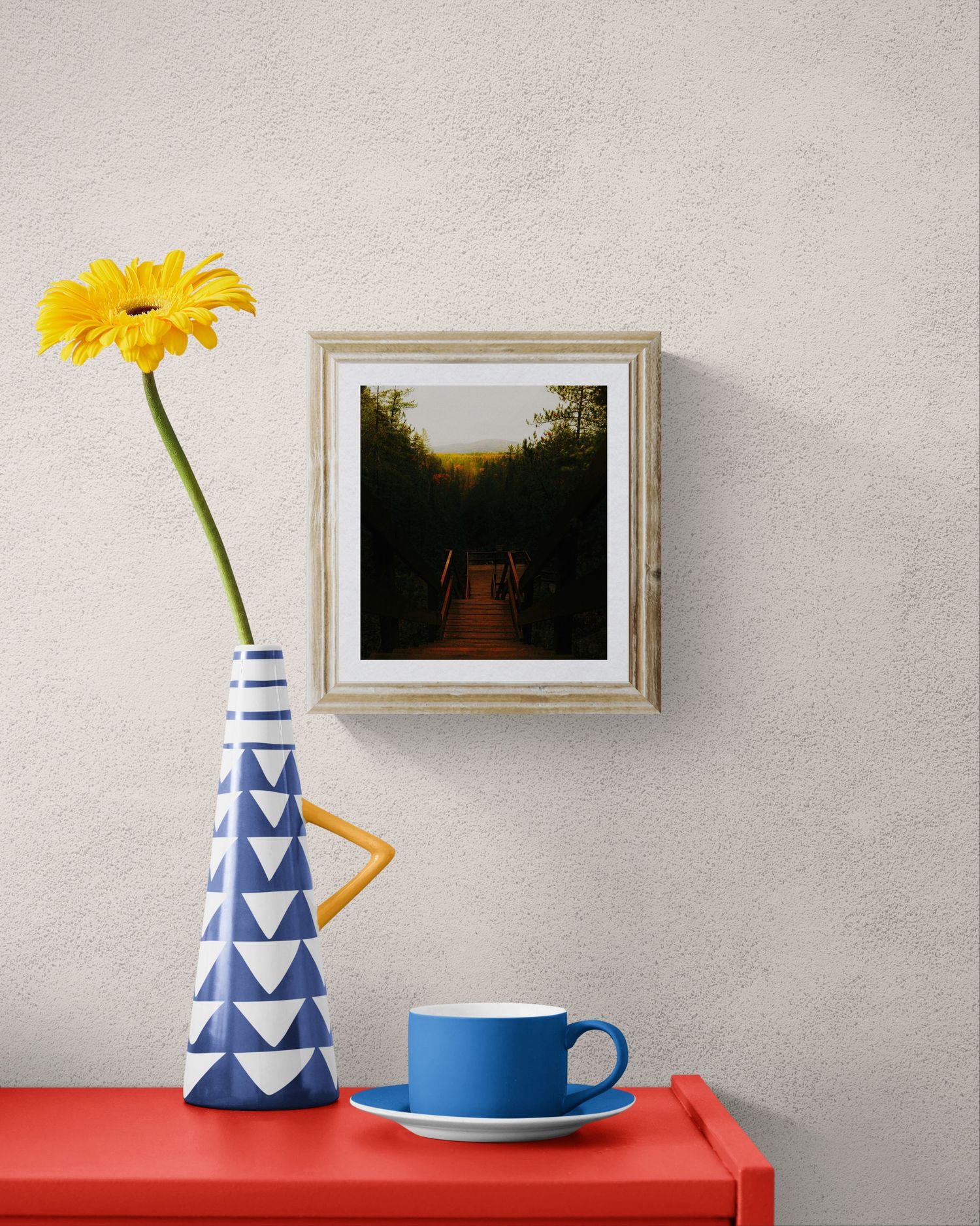 Framed forest path photo on wall above red table with blue cup and striped vase holding yellow gerbera daisy.