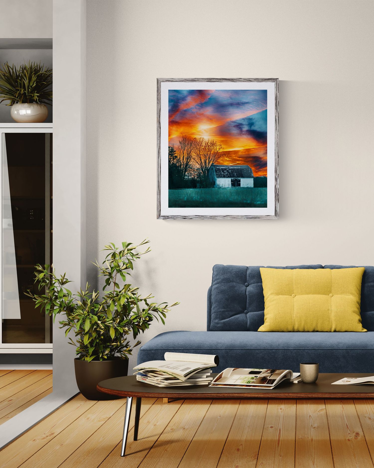 Framed watercolor painting of a cottage at sunset with vibrant orange sky, displayed above a blue sofa in a modern living room.