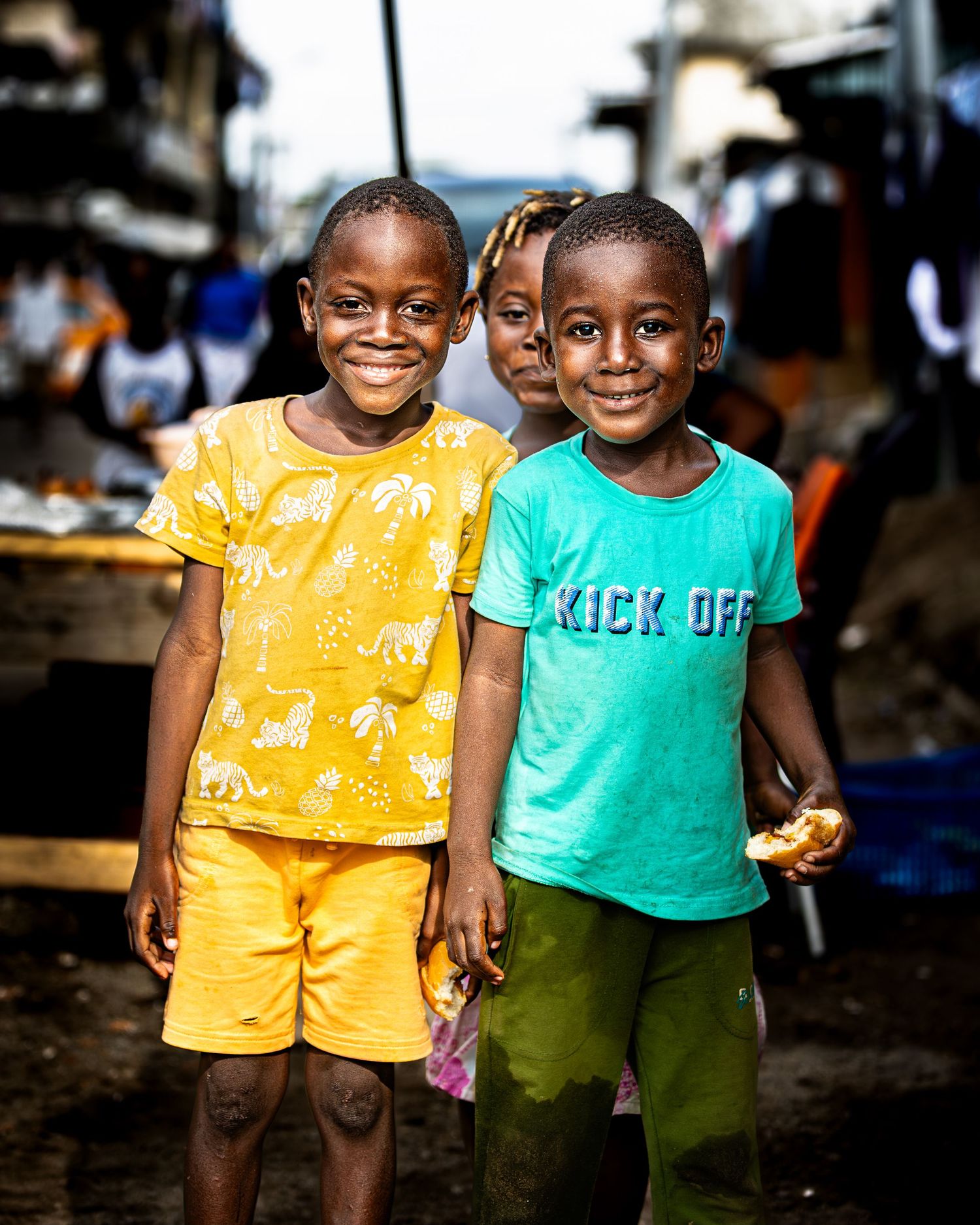 Two children in bright yellow and green outfits smile joyfully while standing together in a market setting.