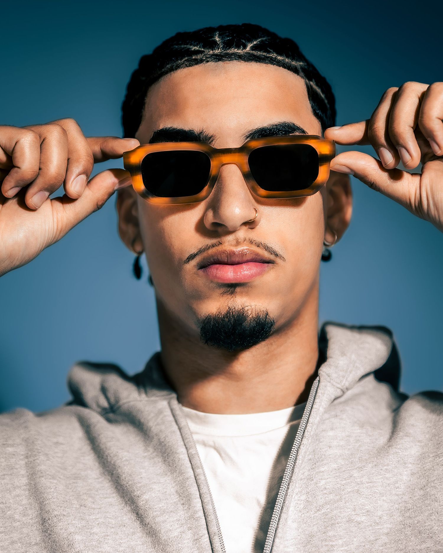 Person in grey hoodie adjusting sunglasses against blue studio backdrop.