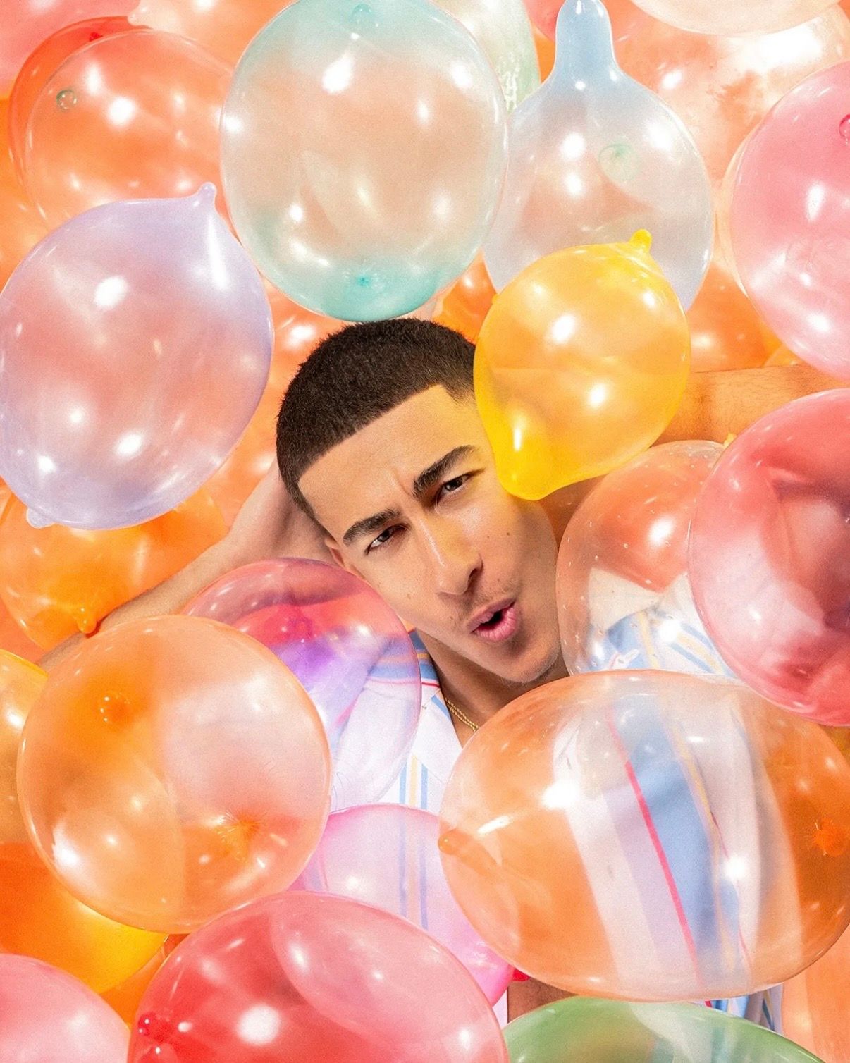 Young man surrounded by colorful pastel balloons making a playful expression in a vibrant orange-toned photo.