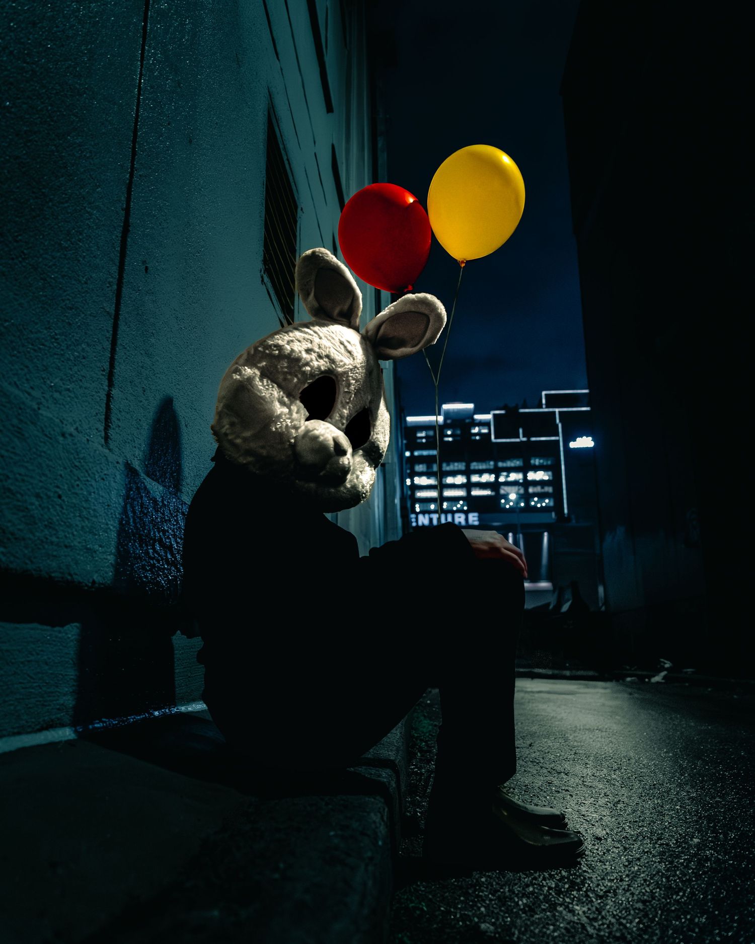 A mysterious figure wearing a bunny mask sits in a dark alley holding red and yellow balloons at night.