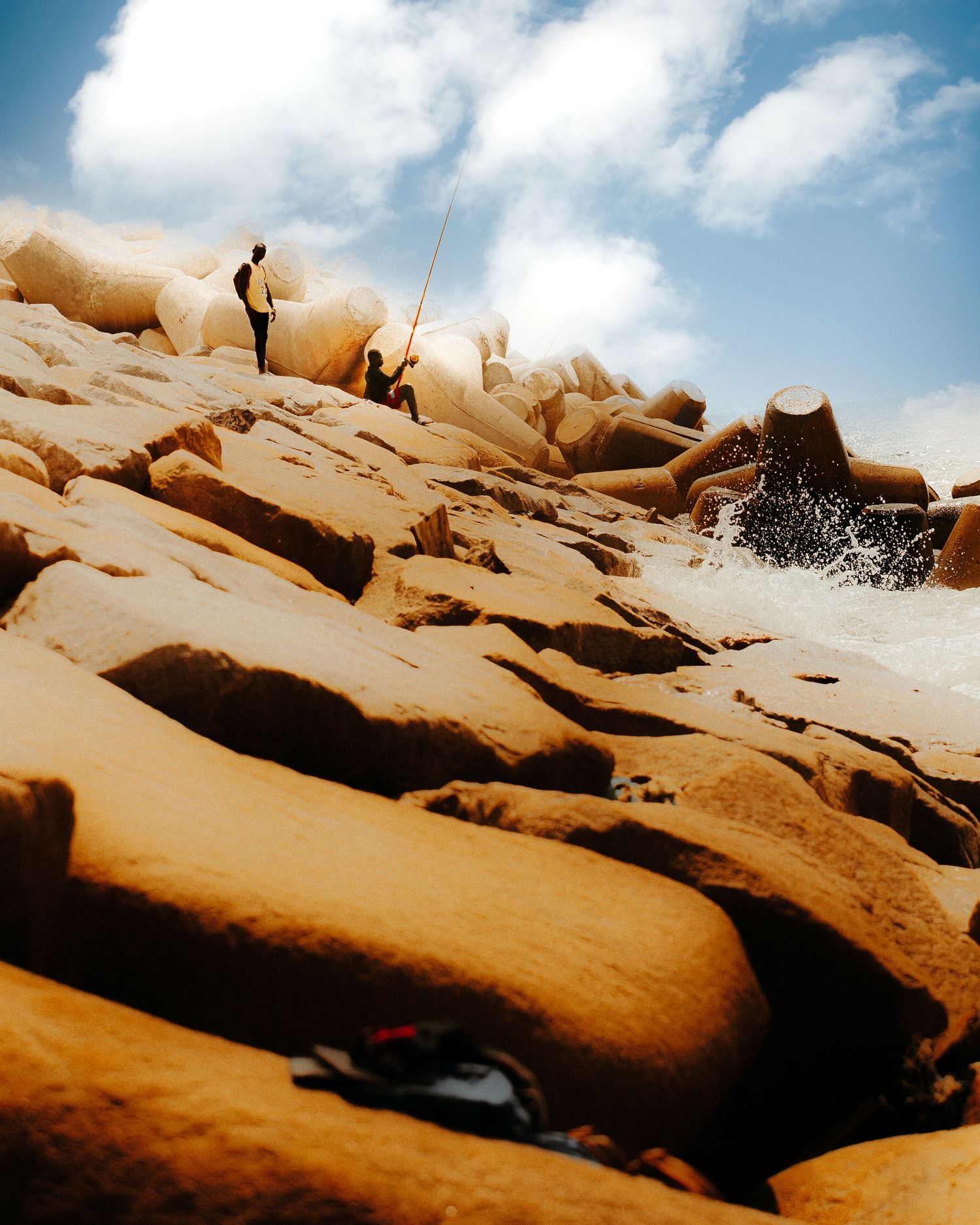 Two people stand on large golden rocks near crashing ocean waves under a partly cloudy sky.