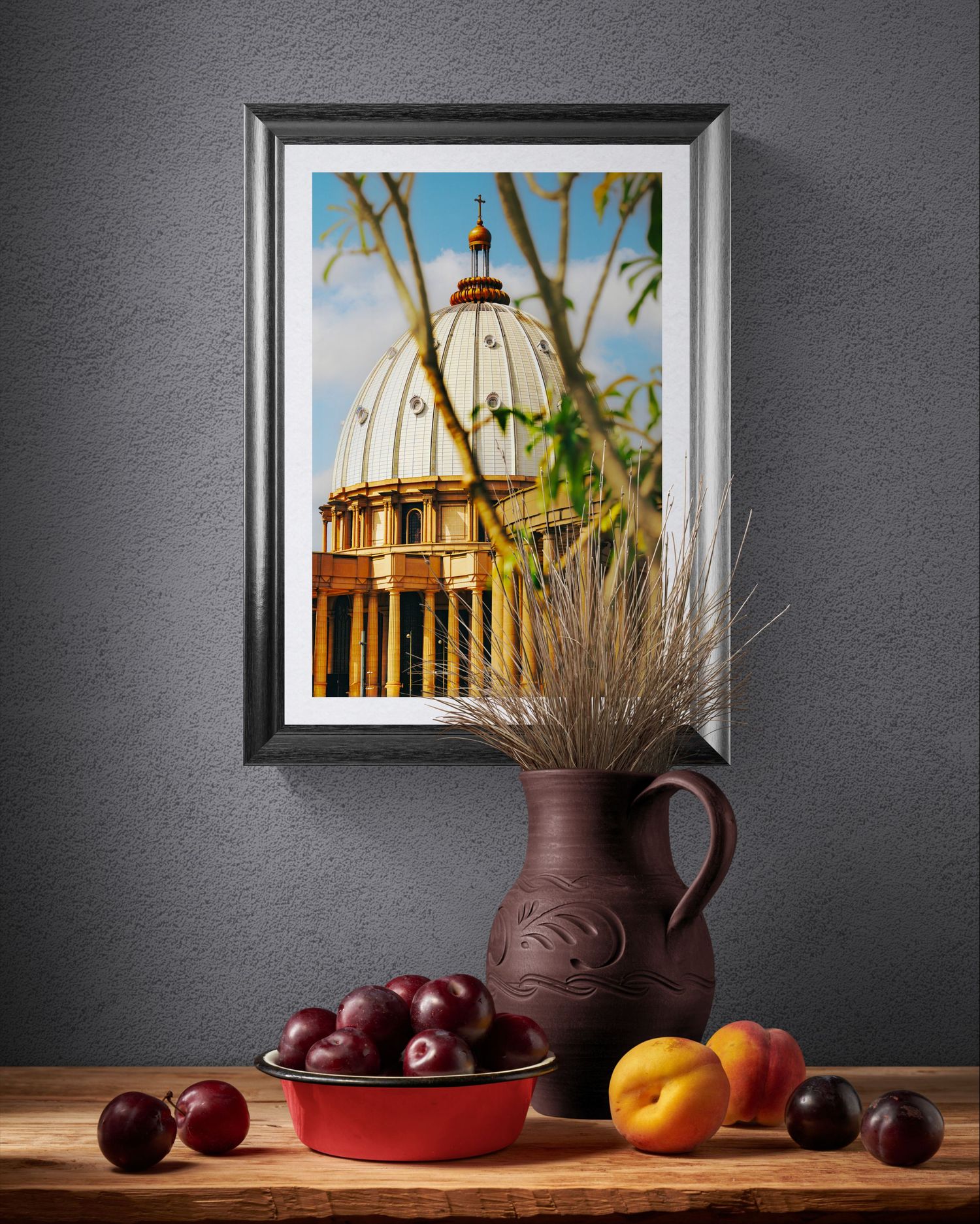 Framed photo of a domed building with bamboo above a wooden table with fruit bowl, plums, peaches, and a brown ceramic jug.