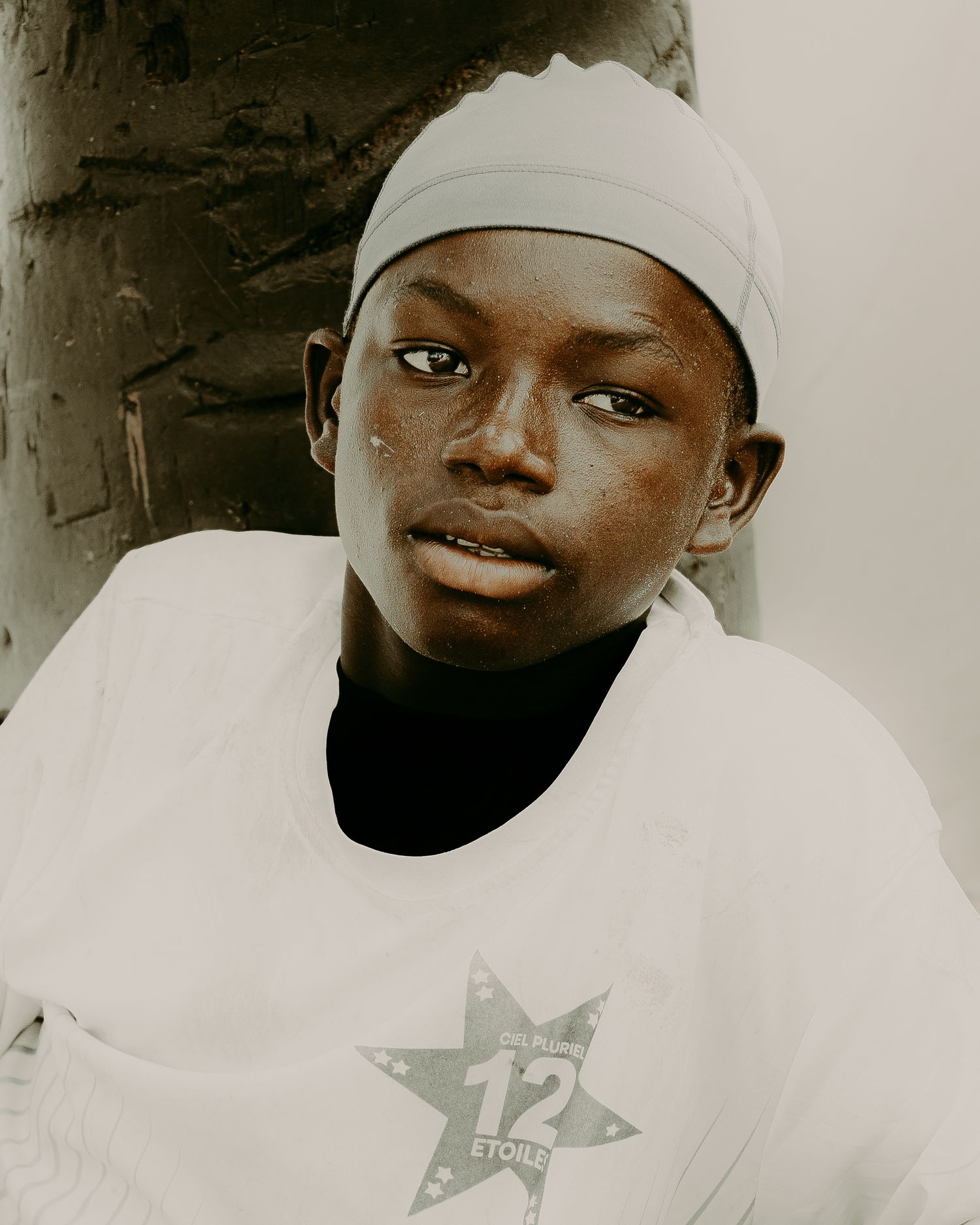 Young person wearing a white 12 Etoiles shirt and light blue head wrap, looking directly at camera.